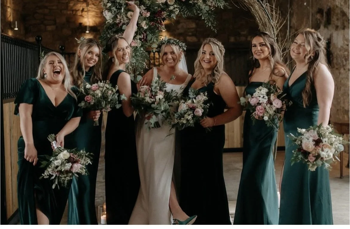 A group of wedding bridesmaids and the bride posing together indoors. The bride is in a white dress, while the bridesmaids are in dark green dresses, holding bouquets of flowers. They are smiling and celebrating.