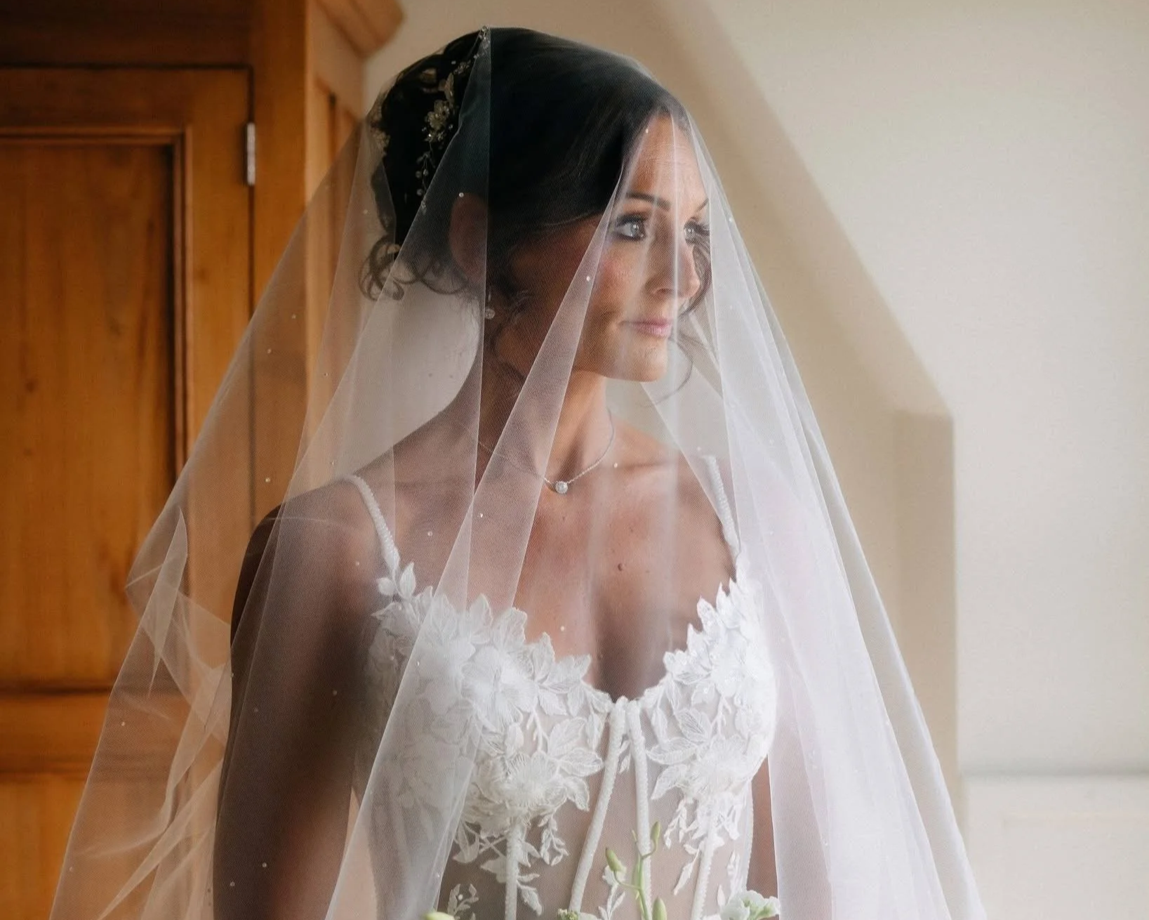 A bride with dark hair, wearing a white wedding dress with floral embroidery, and a veil, looking out the window.