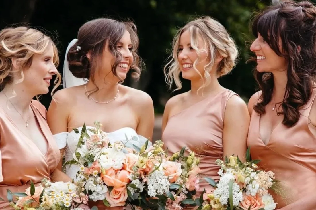 Four women at a wedding, smiling and looking at each other, holding bouquets of pink and white flowers.