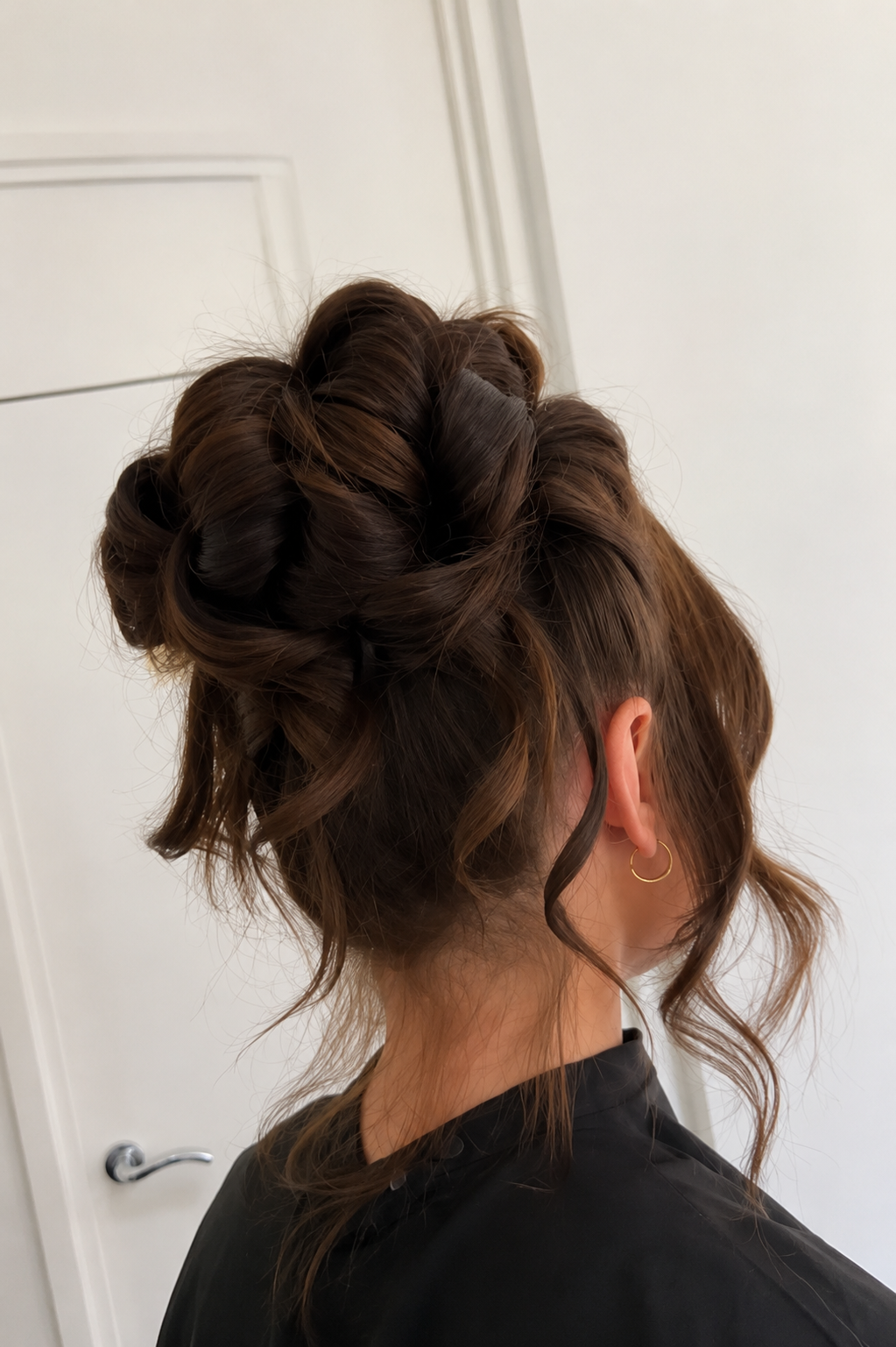 A woman with medium brown wavy hair styled in an elaborate updo, with loose tendrils framing her face. She is wearing small gold hoop earrings and a black top, standing in front of a white door.