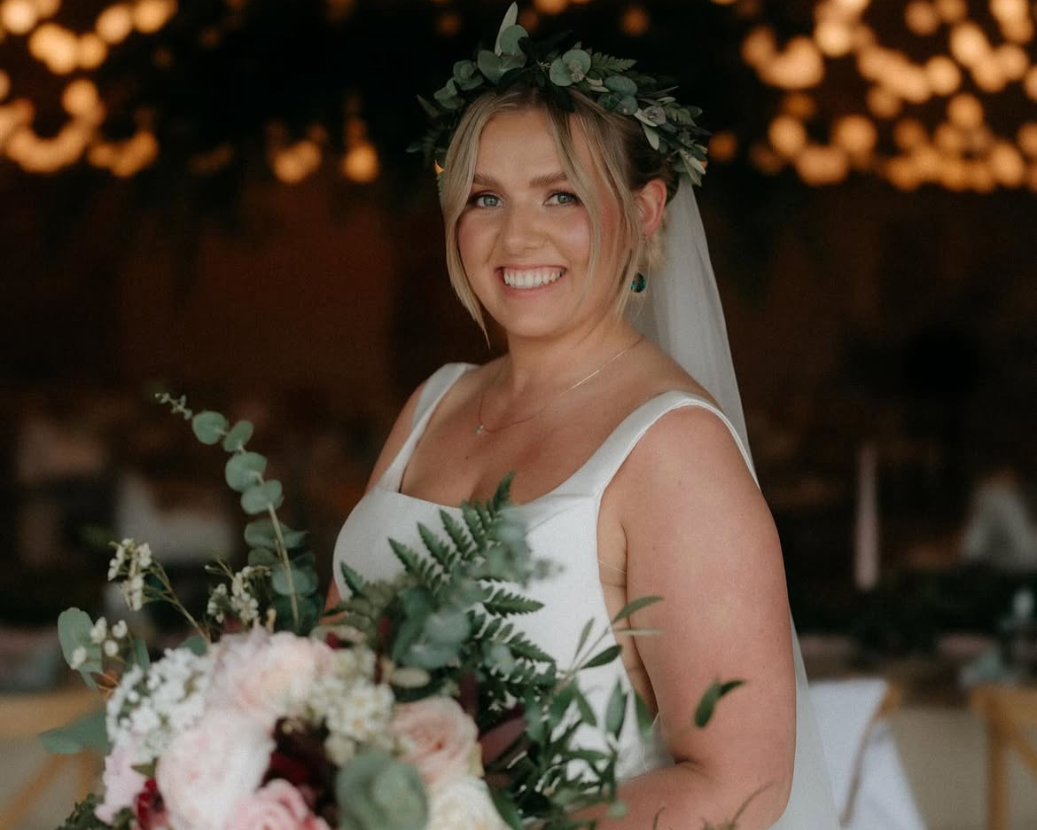 A smiling bride with blonde hair wearing a white wedding dress, a floral crown, and holding a bouquet of flowers and greenery at her wedding reception.