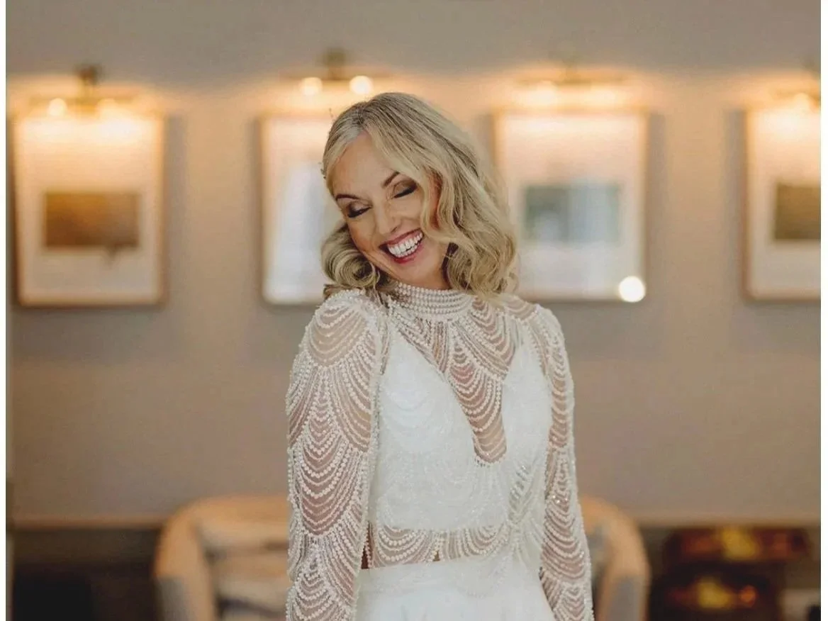 A woman with blonde hair smiling and looking down, wearing a white dress with intricate beaded details, inside a room with framed pictures on the wall and warm lighting.
