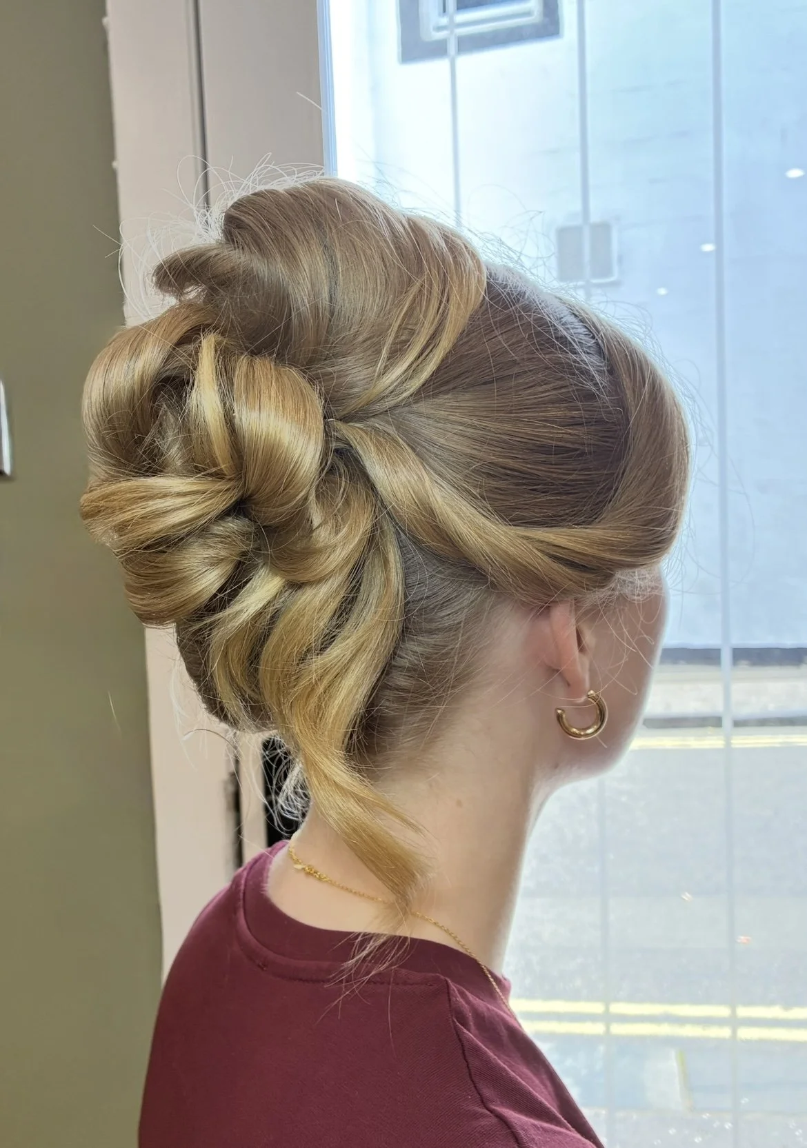 A woman with blonde hair styled in an elegant updo with curls, wearing gold hoop earrings and a maroon top, standing by a window with natural light.