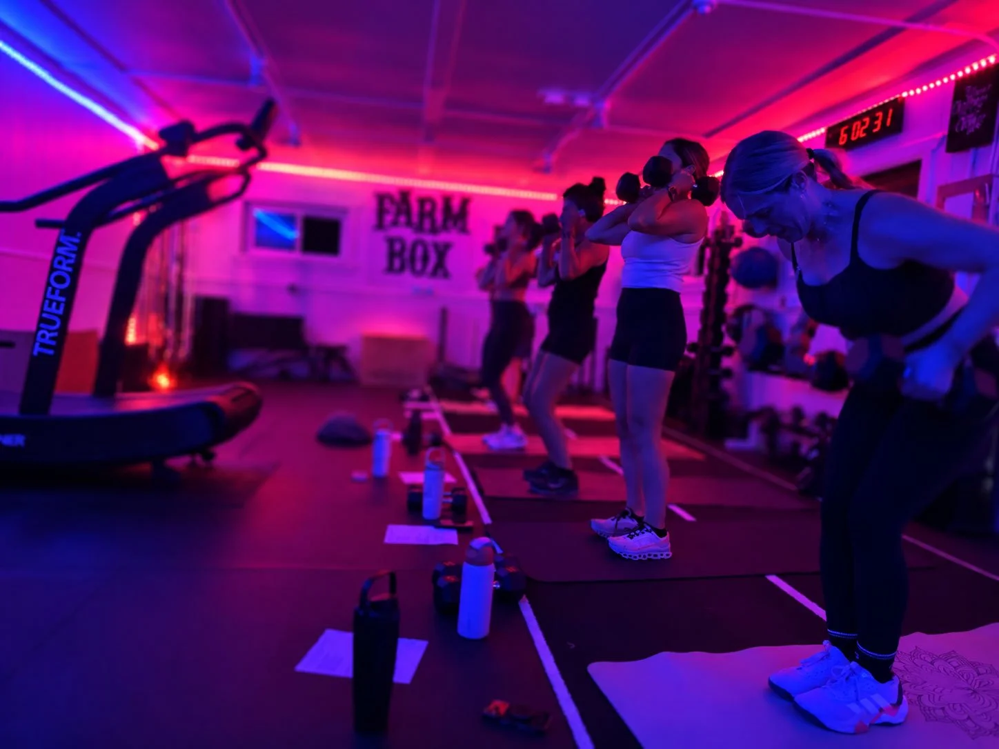 Women doing weightlifting exercises in a dimly lit gym with pink and purple lighting, with water bottles and workout mats on the floor.