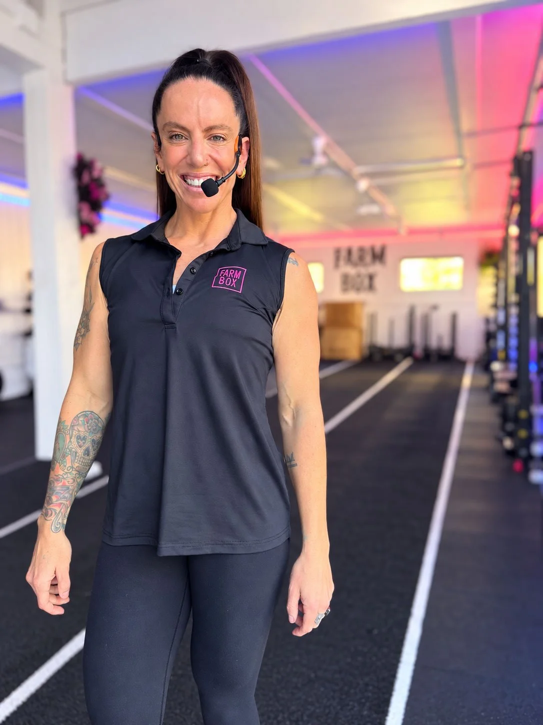 A woman with tattoos on her right arm, wearing a black sleeveless polo shirt with a pink 'FARM BOX' logo and a headset, smiling inside a gym with colorful LED ceiling lights.