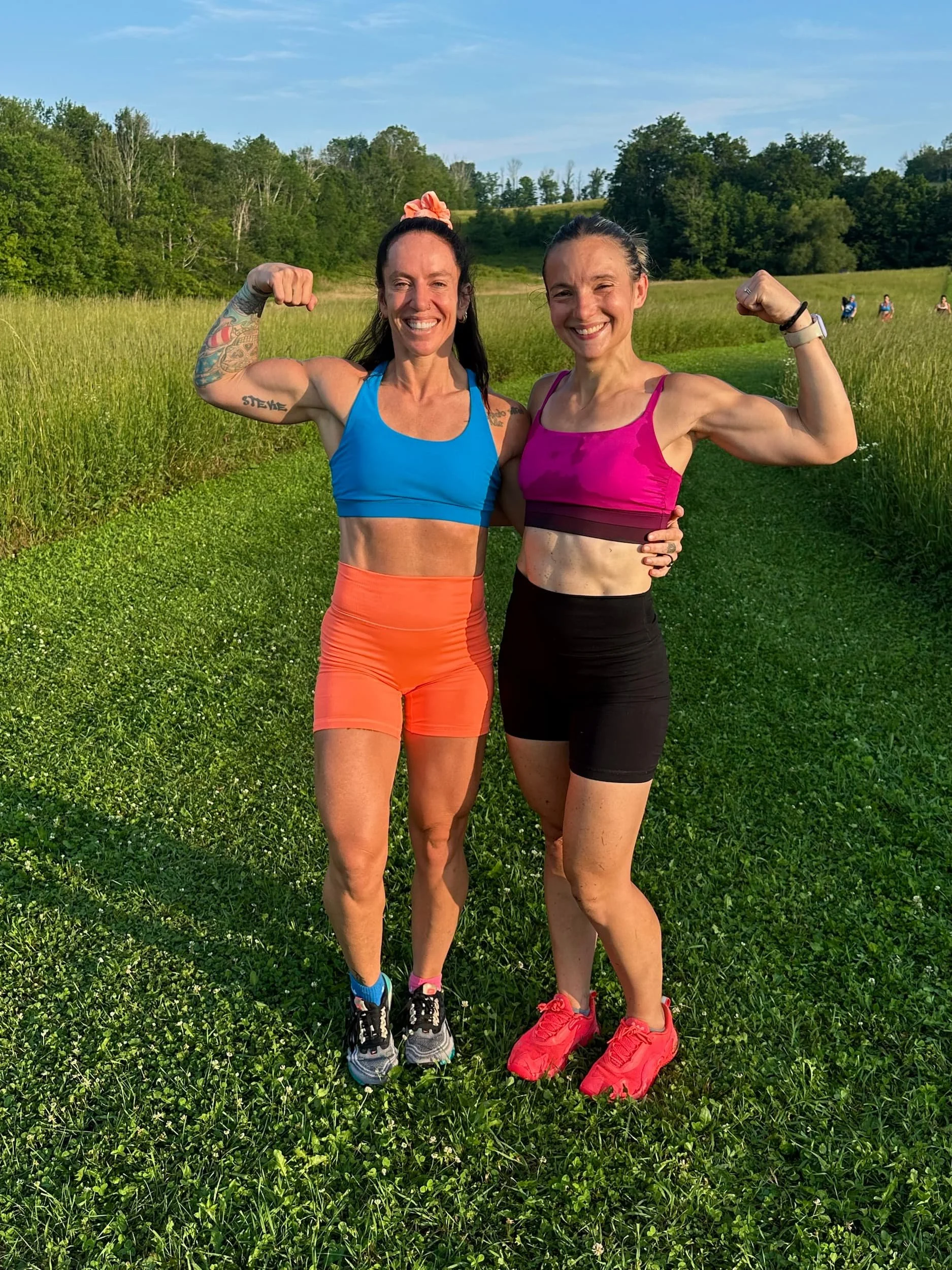 Two women in athletic wear flexing their biceps and smiling on a grassy field with trees and a blue sky in the background.