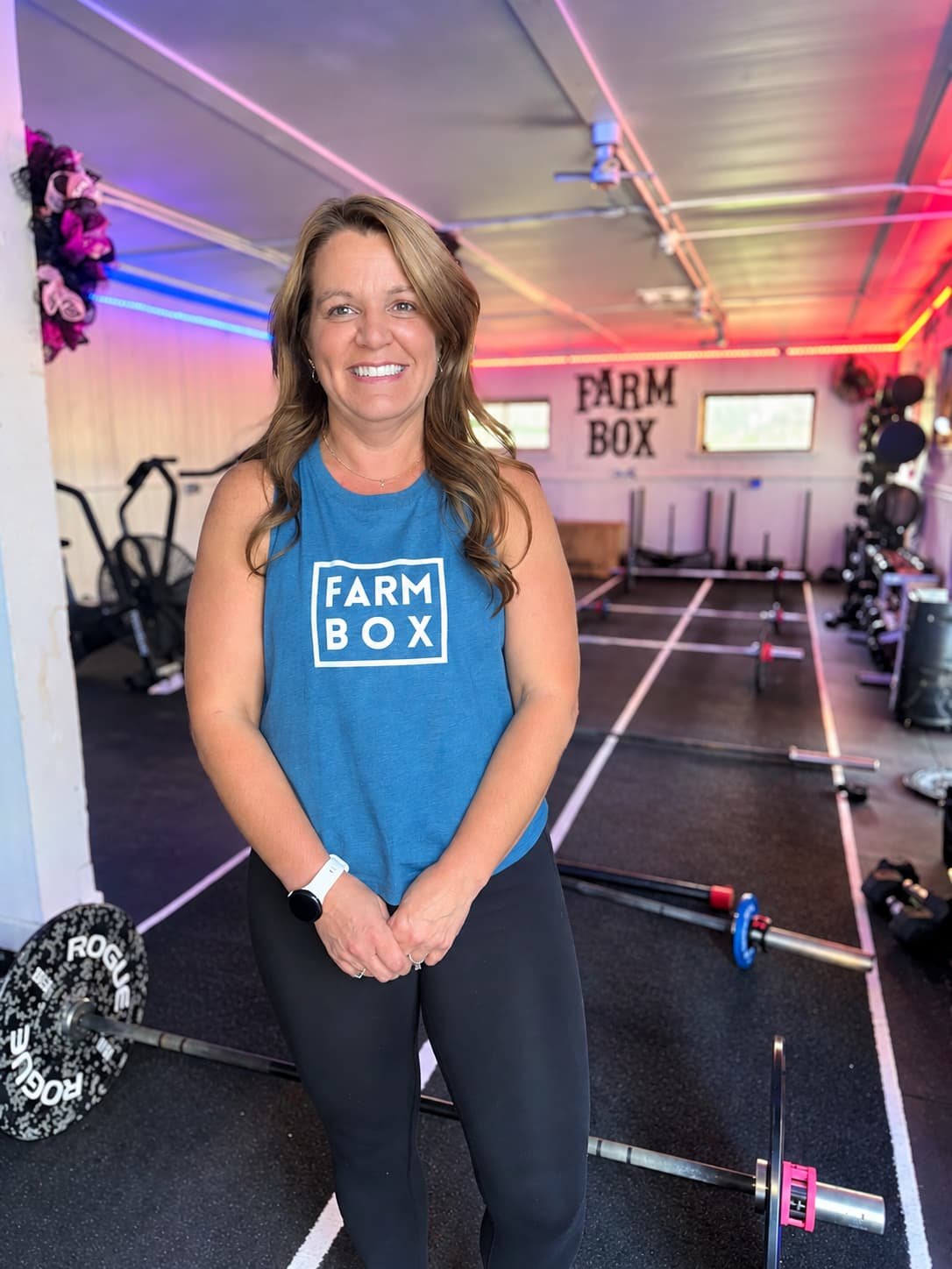 Woman in a blue sleeveless shirt with 'FARM BOX' logo, standing in a gym with weightlifting equipment, weight plates, and barbells, and a sign that says 'FARM BOX' on the wall.