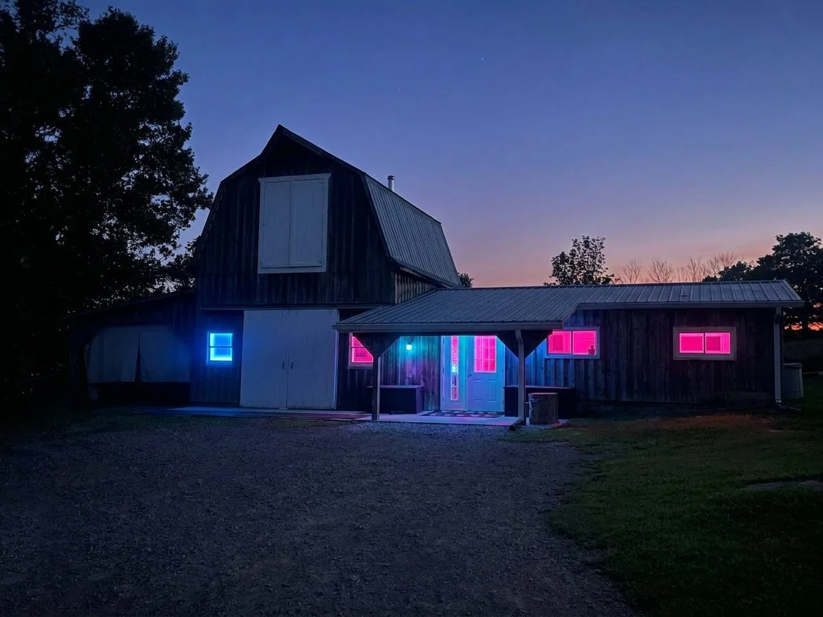 A barn with neon pink and blue lights illuminating the windows and doorway at dusk, with a gravel pathway leading up to it and trees in the background.