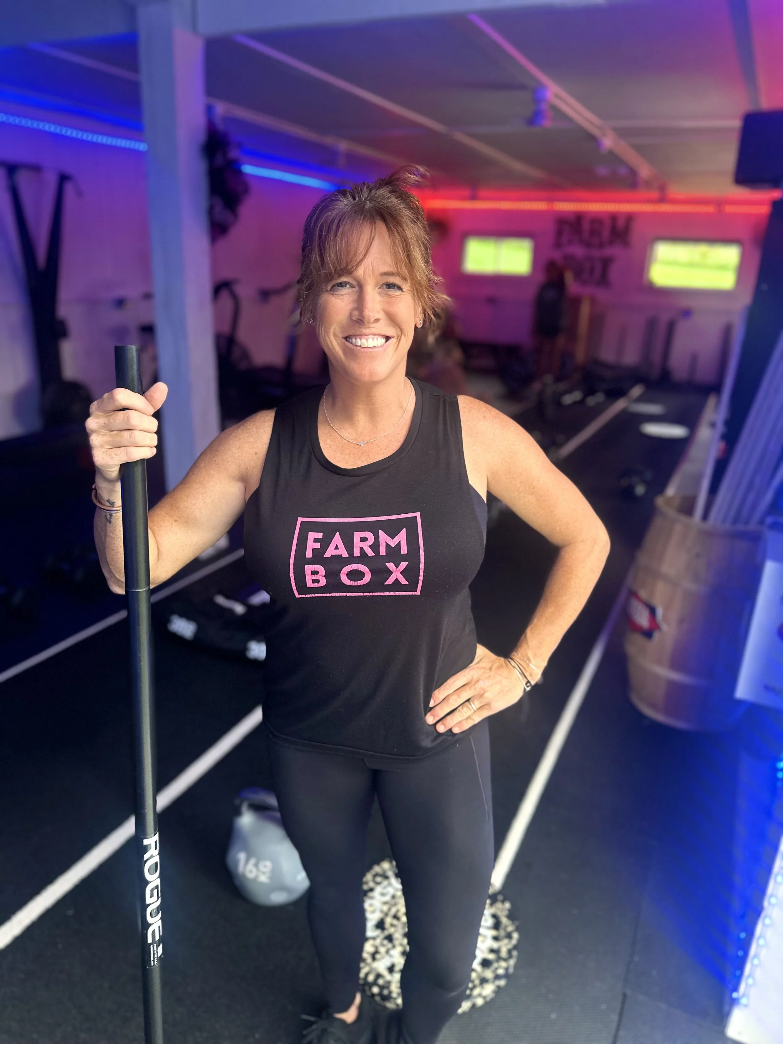 A smiling woman standing in a gym holding a black exercise pole, with colorful neon lighting and fitness equipment in the background.