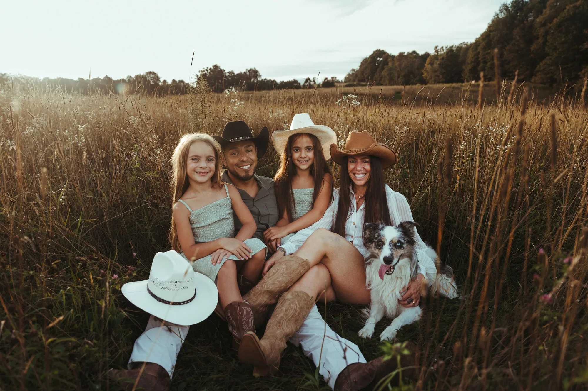 A family of four and a dog sitting together in a grassy field with trees in the background during sunset.