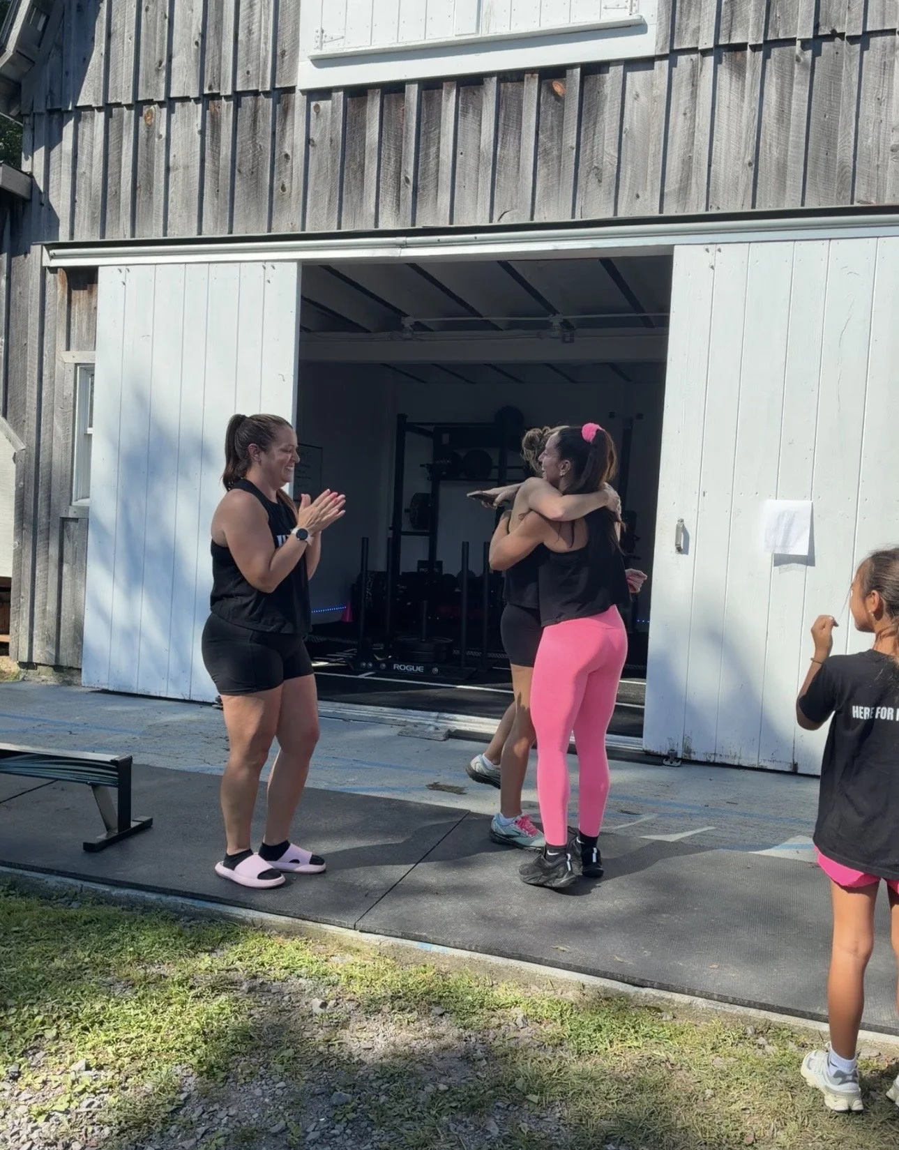 Women and children outside a gym, celebrating and hugging after a workout session. One woman claps while others cheer and smile on a paved area near a barn-like building.