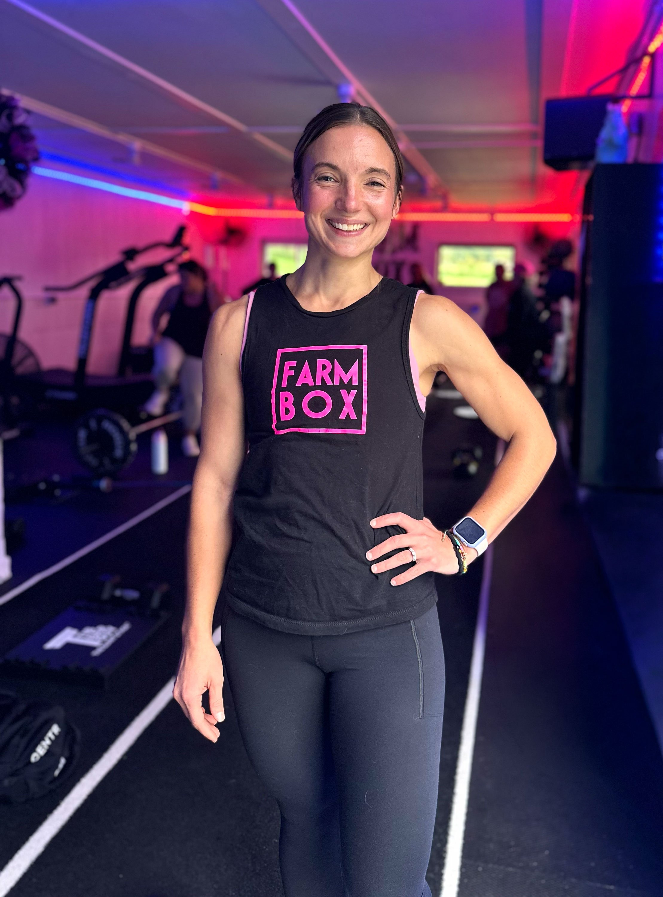 A smiling woman in workout clothes standing inside a gym with colorful neon lighting, with fitness equipment and exercise machines in the background.