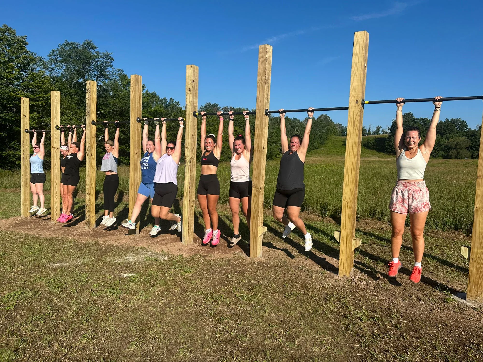 Group of women hanging from a horizontal bar on an outdoor obstacle course with a wooded background under a blue sky.