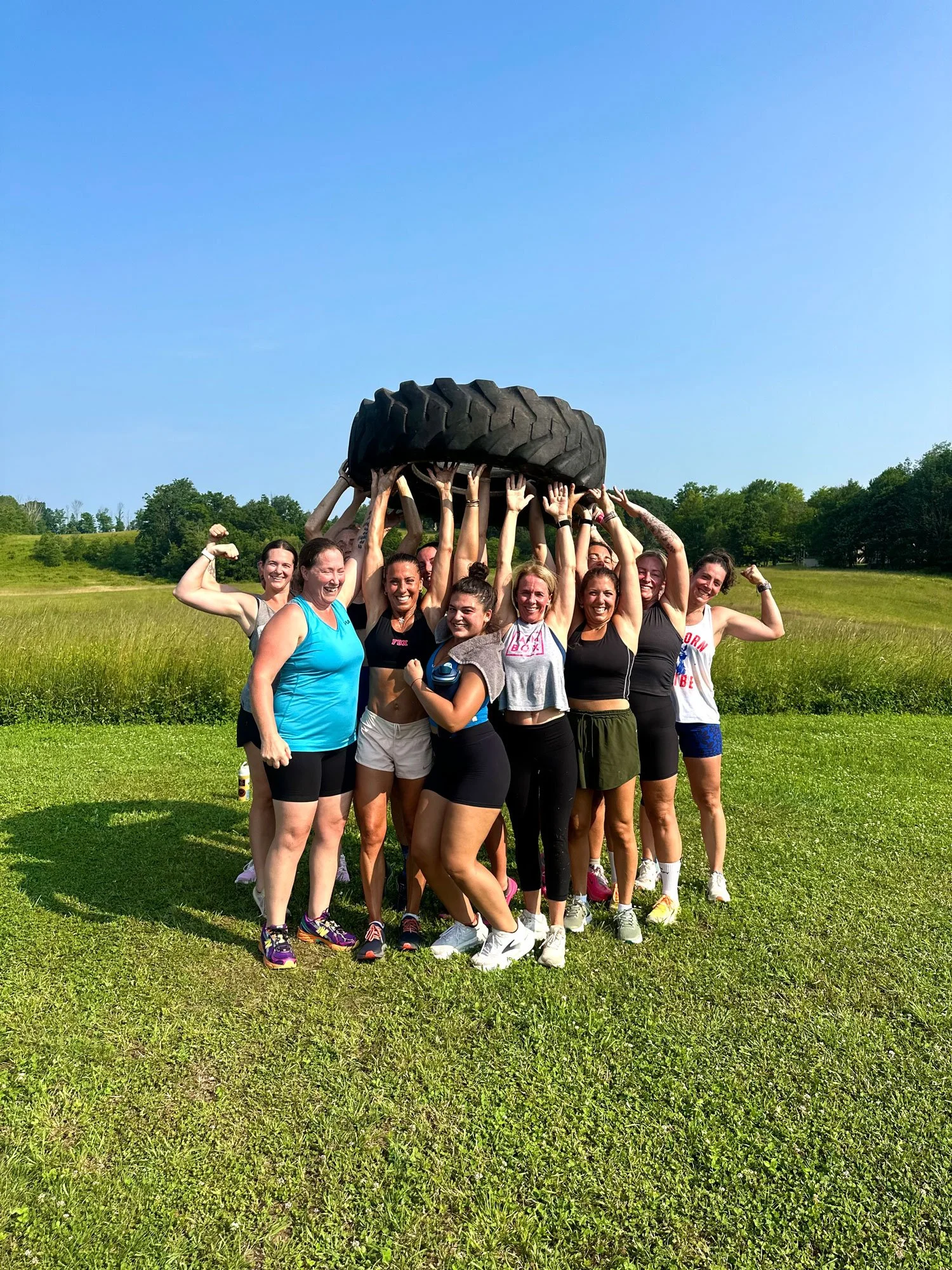A group of women in athletic clothing lifting a large tire together outdoors in a grassy field on a sunny day.