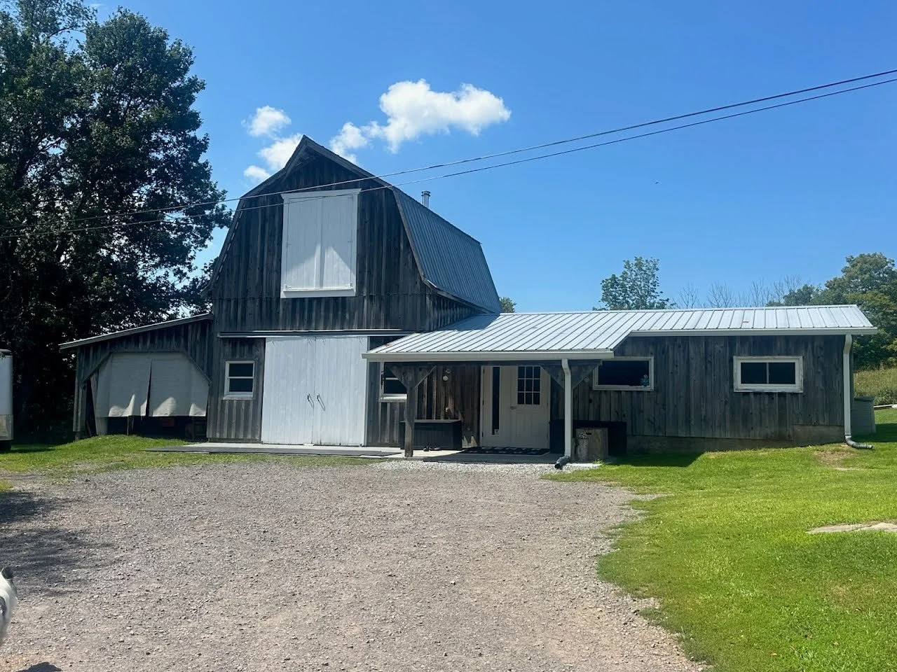 A rustic barn with weathered wooden exterior, metal roof, small windows, and a small covered porch, set in a rural area with gravel driveway, green grass, trees, and a blue sky with a few clouds.