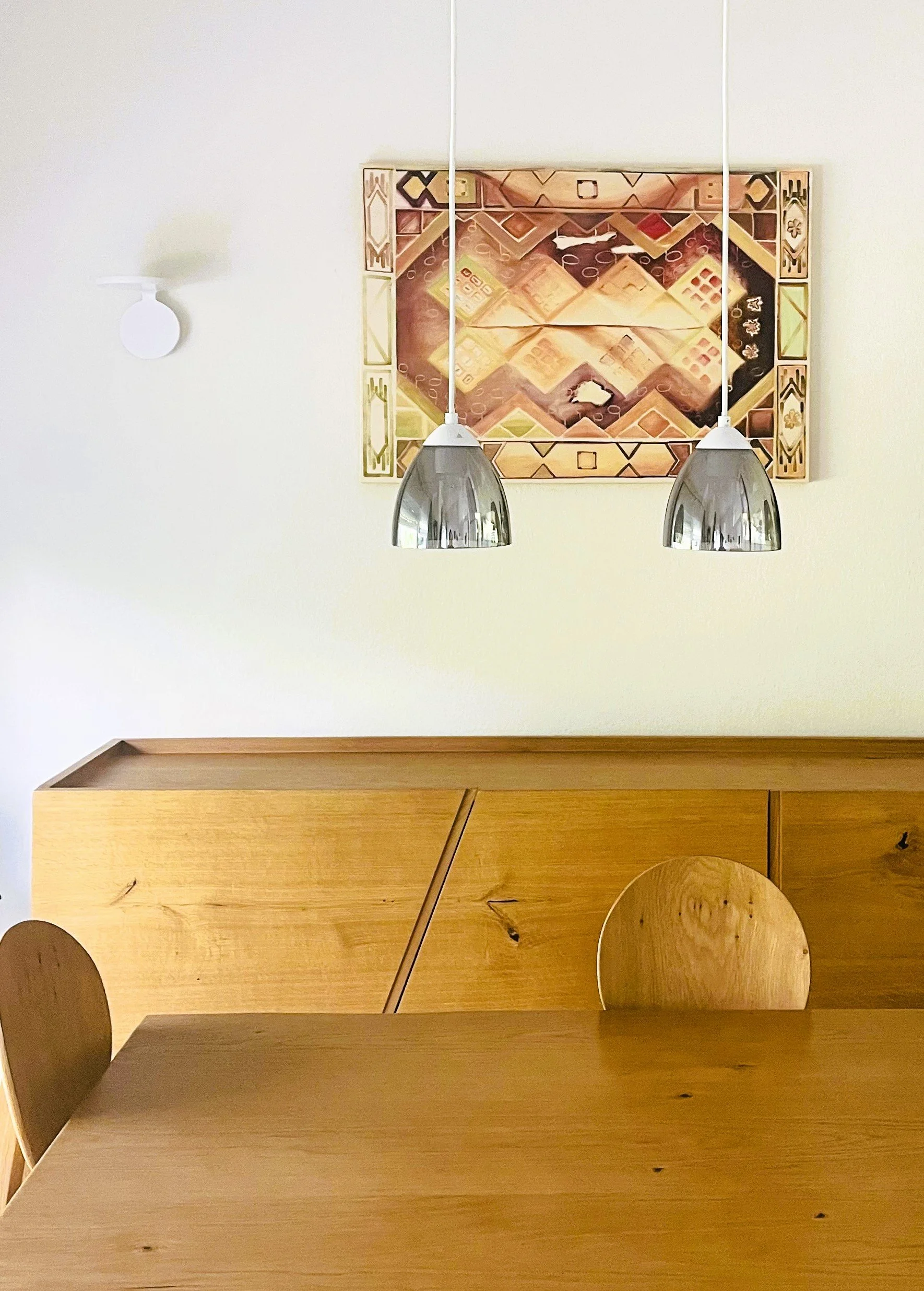 Two metal pendant lights hang above a wooden shelf or table, with a decorative framed artwork and a small white wall lamp on a plain cream-colored wall.