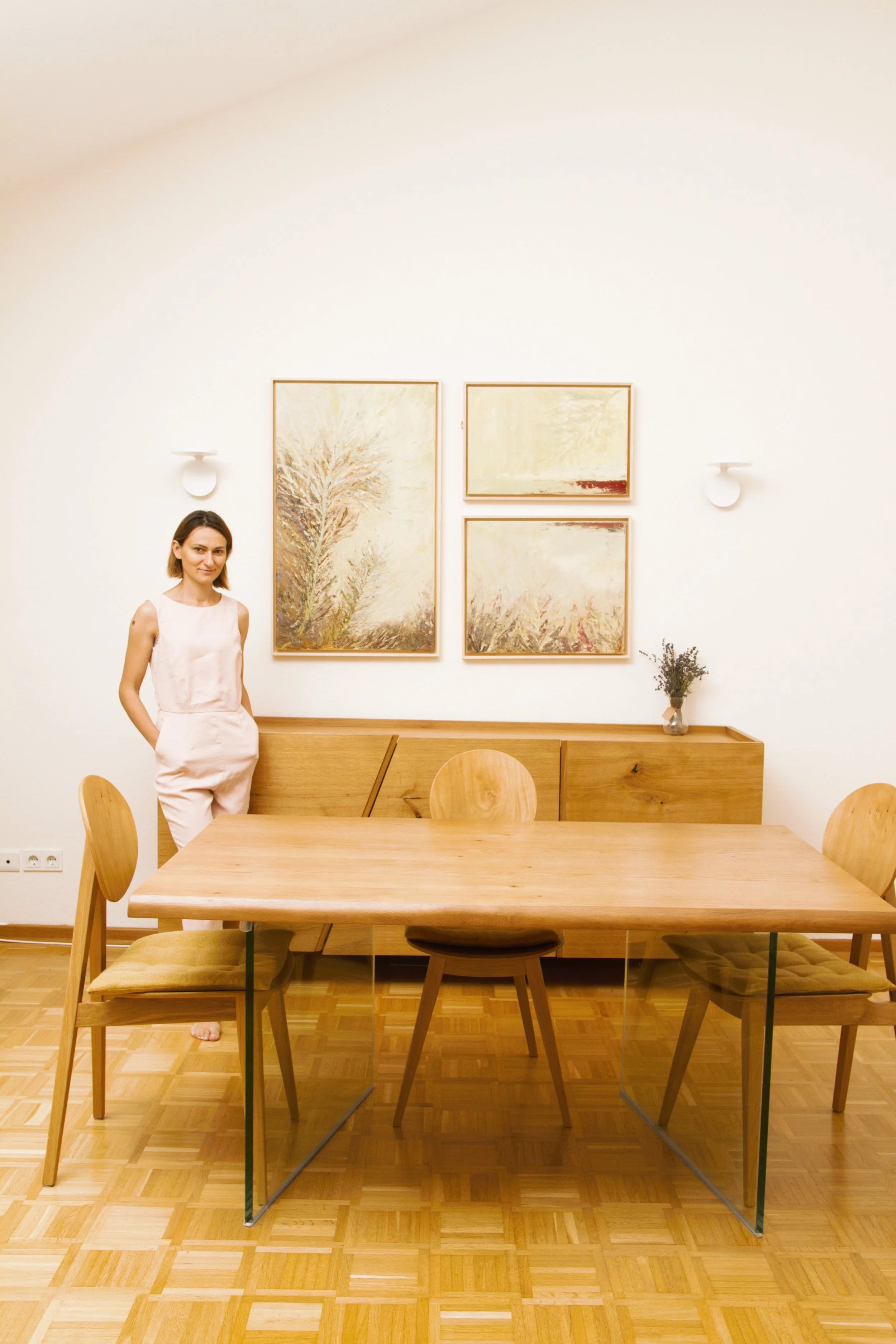 A woman standing next to a wooden dining table in a room with white walls, four framed abstract art pieces, and a small vase with lavender on a sideboard.