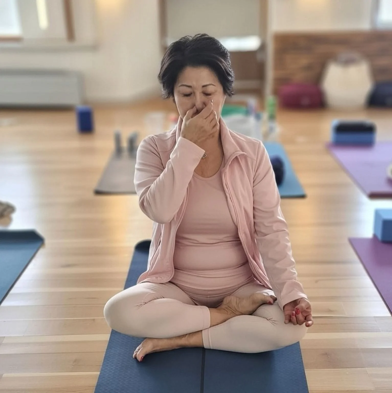 Une femme pratiquant la méditation dans une salle lumineuse, assise en position du lotus sur un tapis de yoga, portant des vêtements roses et silencieusement concentrée.