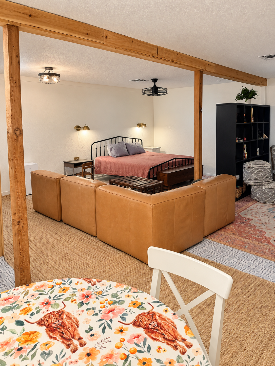 Living room with a beige sectional sofa, a black metal bed with pink bedding, wooden beams, and various decor items including a patterned rug and a bookshelf.