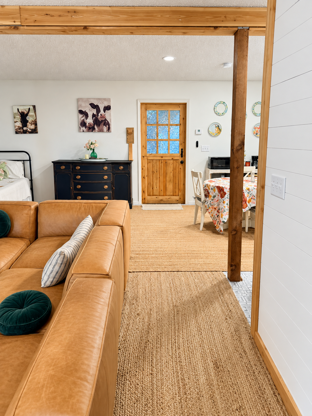 Interior view of a cozy living space featuring a tan leather sofa with decorative pillows, a black dresser with vases and flowers, a wooden door with decorative window, a dining area with a round table and chairs covered with a colorful floral tablec