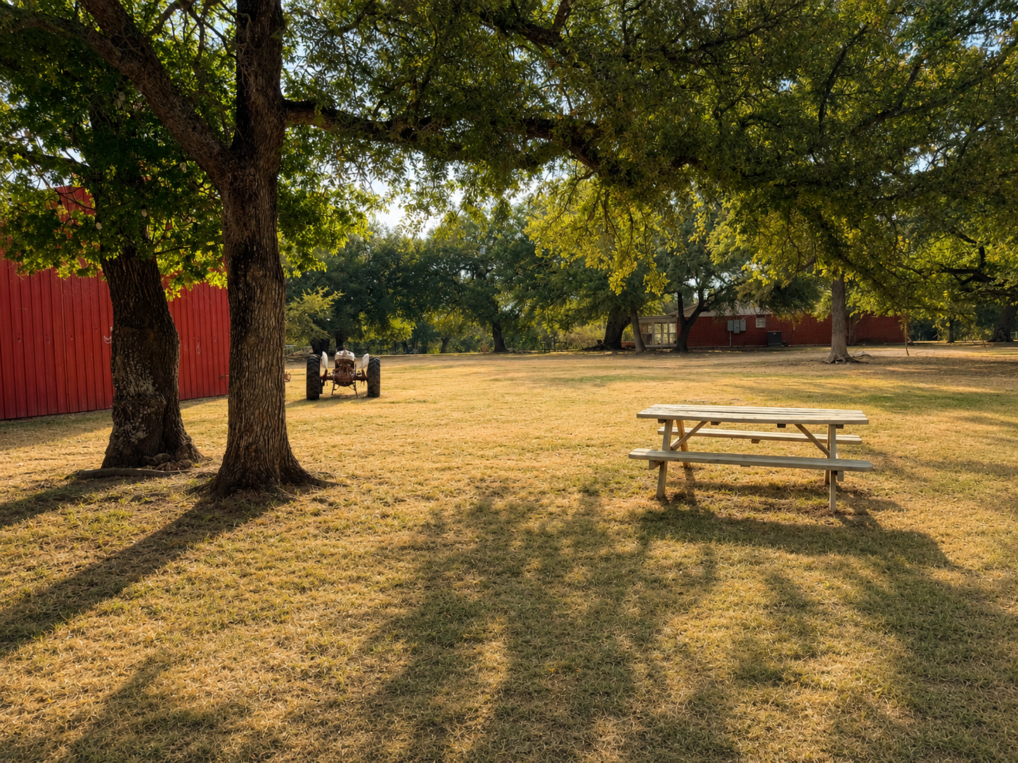 A rural outdoor scene with a grassy area shaded by large trees; a picnic table is in the foreground, and a vintage tractor is near a red barn on the left side of the image.