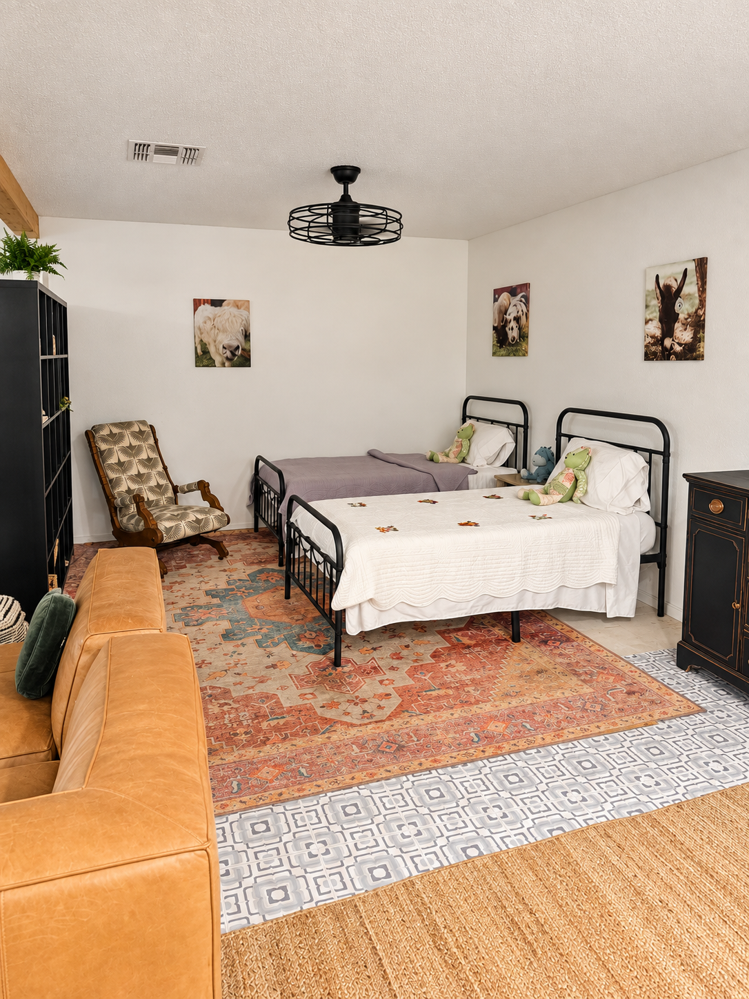 A cozy bedroom with two black metal beds, decorated with stuffed animals and pillows, featuring a patterned rug, wooden furniture, and wall art of farm animals.
