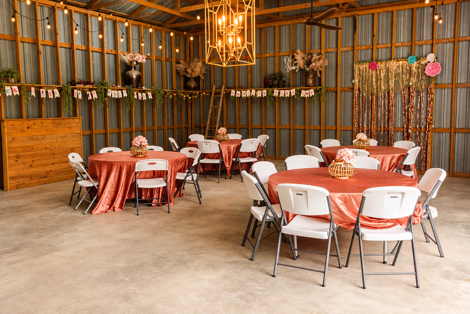 Decorated event space with round tables covered in coral-colored tablecloths, each with a floral centerpiece, white chairs, string lights, and a decorated backdrop, likely for a celebration or party.