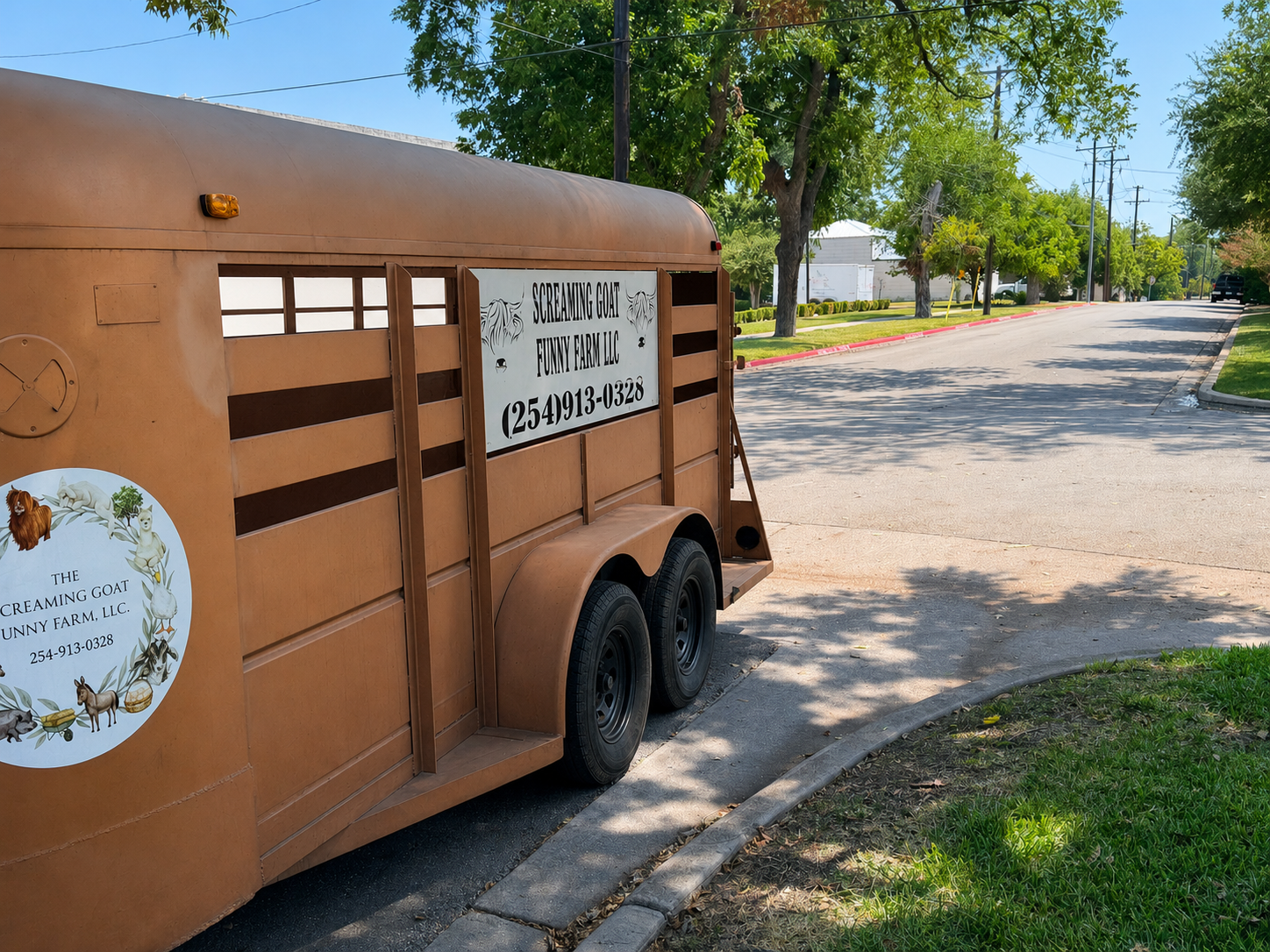 A mobile animal shelter trailer parked beside a tree-lined street on a sunny day, with signs indicating it is for 'Screaming Goat Funny Farm LLC' and displaying a phone number.