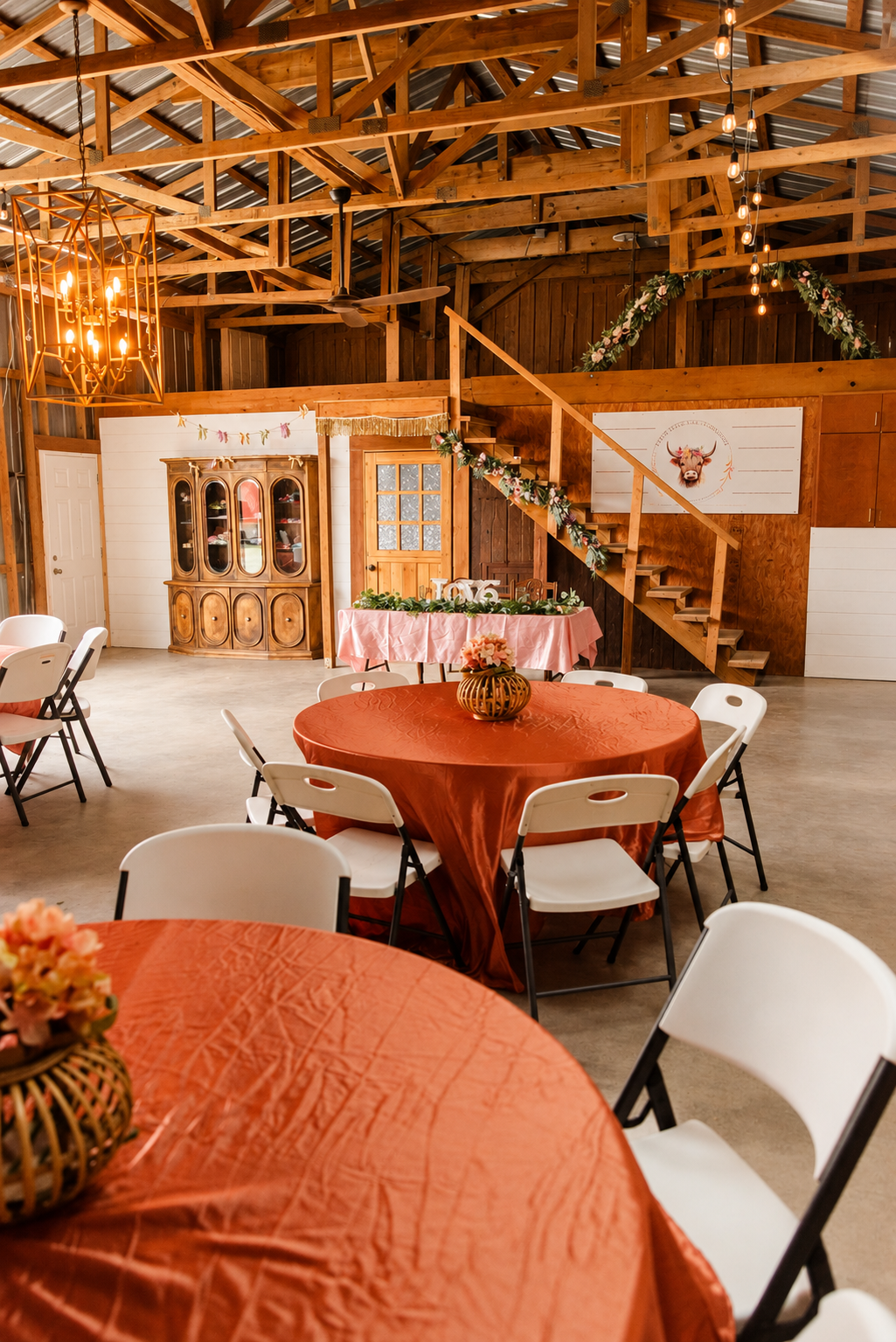 Interior of a rustic barn decorated for a celebration, featuring round tables with orange tablecloths, white chairs, floral centerpieces, a wooden staircase adorned with garland, a wooden cabinet, and a painted deer head mural on the wall.