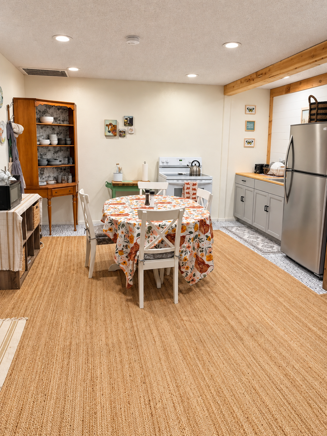 A cozy kitchen with a round dining table covered with a floral tablecloth, surrounded by four white chairs. There is a wooden china cabinet on the left, a stove in the back, and light gray cabinets on the right. The floor is covered with a tan rug, a