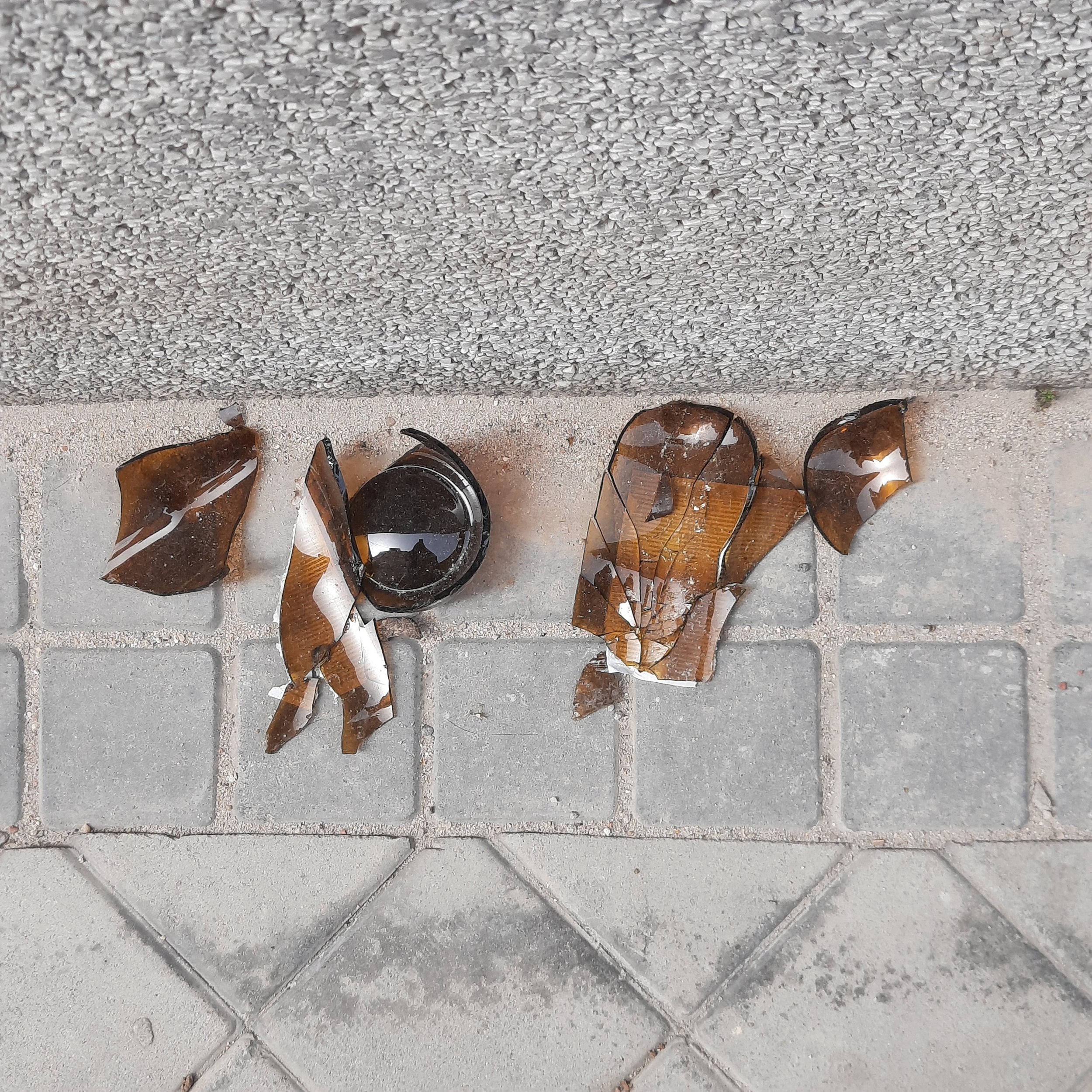Broken amber-colored glass bottles lying on a concrete sidewalk with gray tiling in the background.