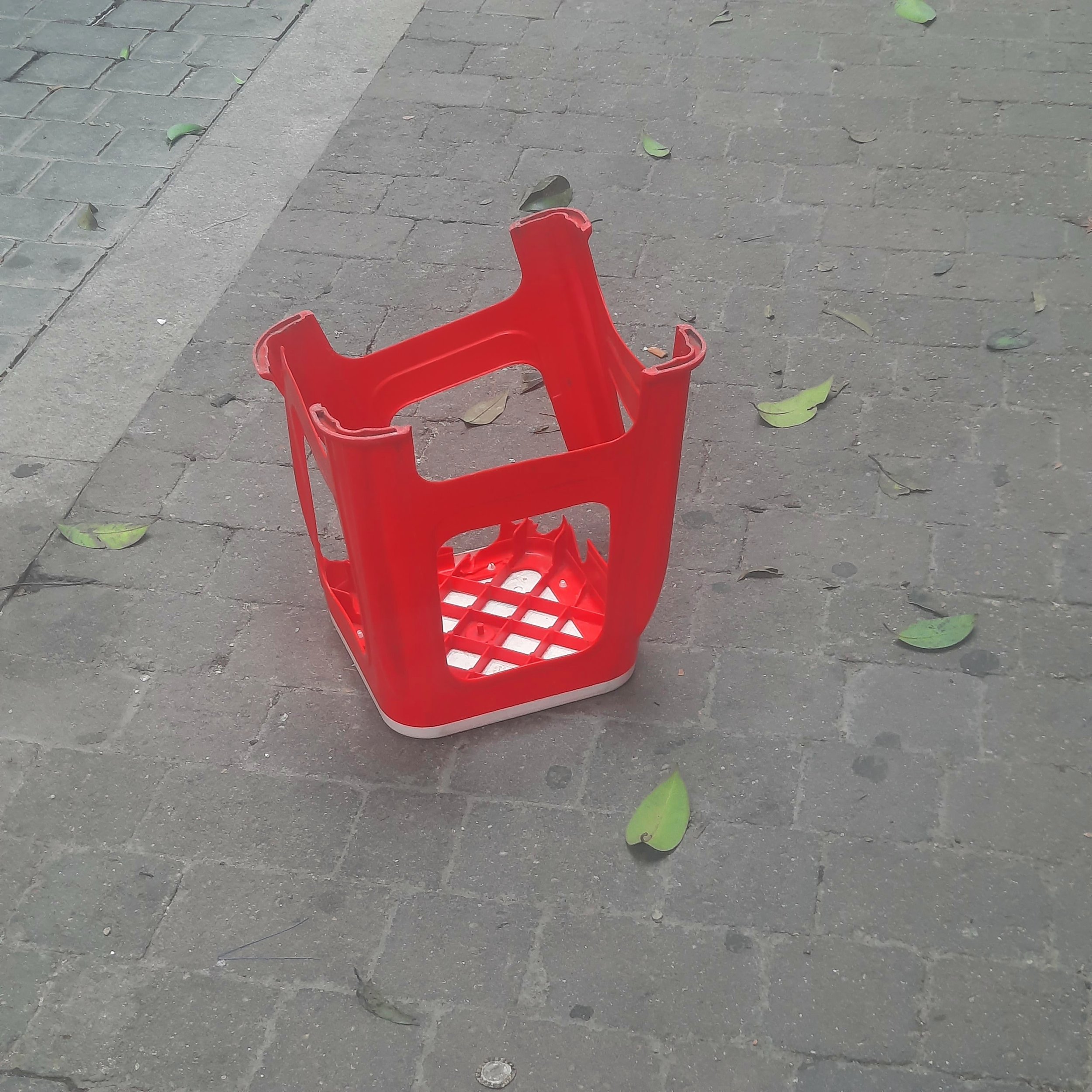 A red plastic chair upside down on a sidewalk with scattered green leaves and cars partially visible in the background.
