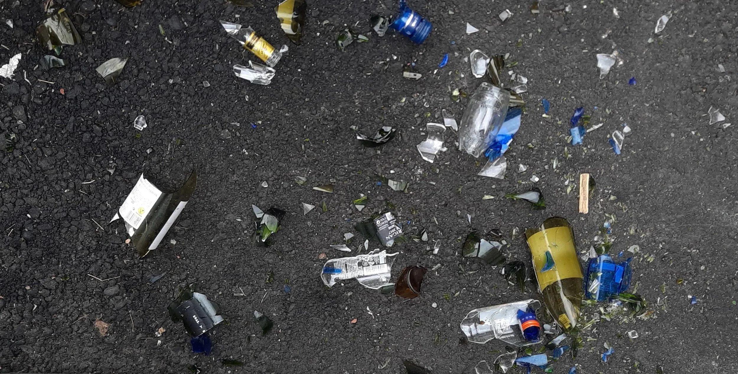 Scattered trash on asphalt surface, including broken glass bottles, cans, and debris.