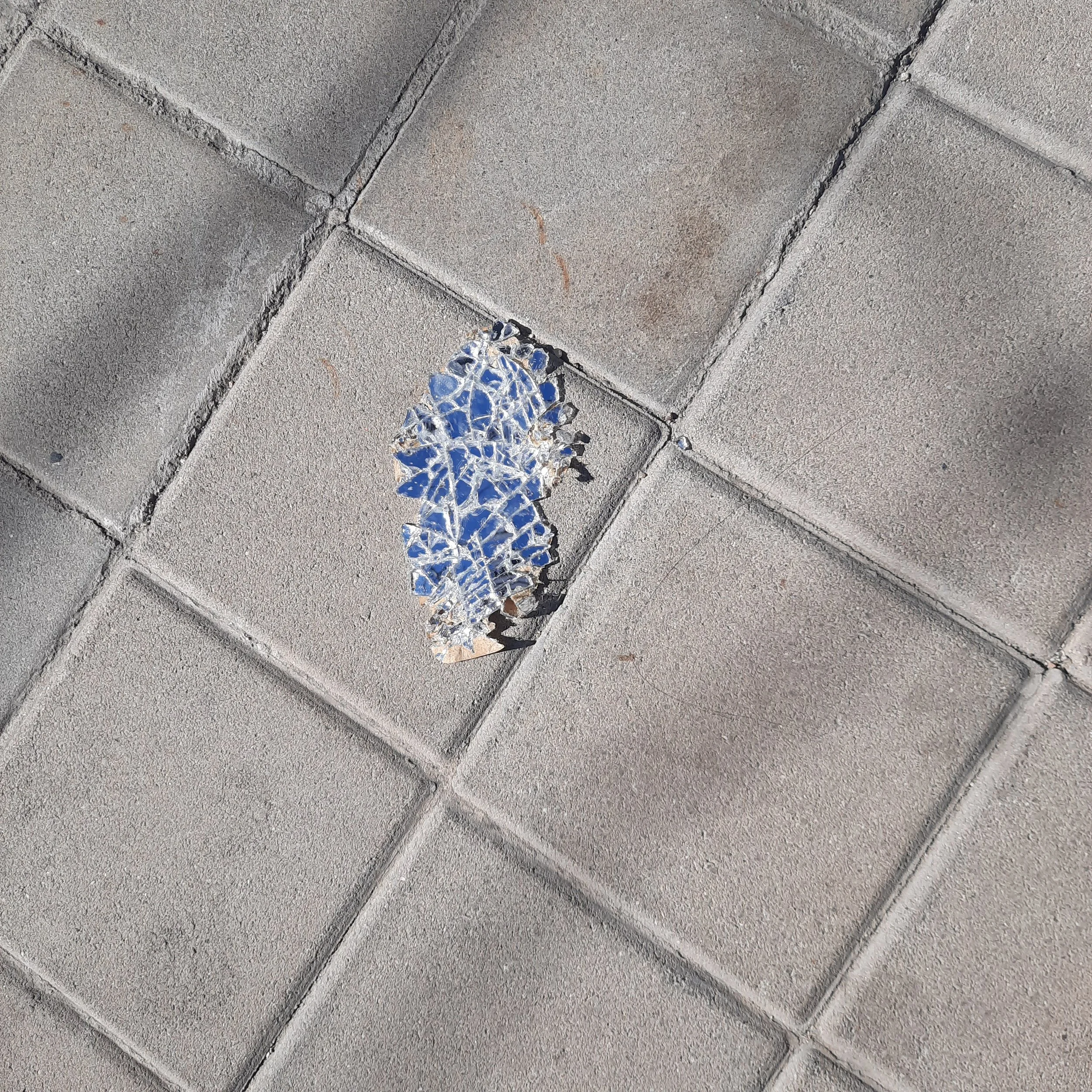 Cracked and damaged concrete sidewalk with a large area of broken glass embedded in it.