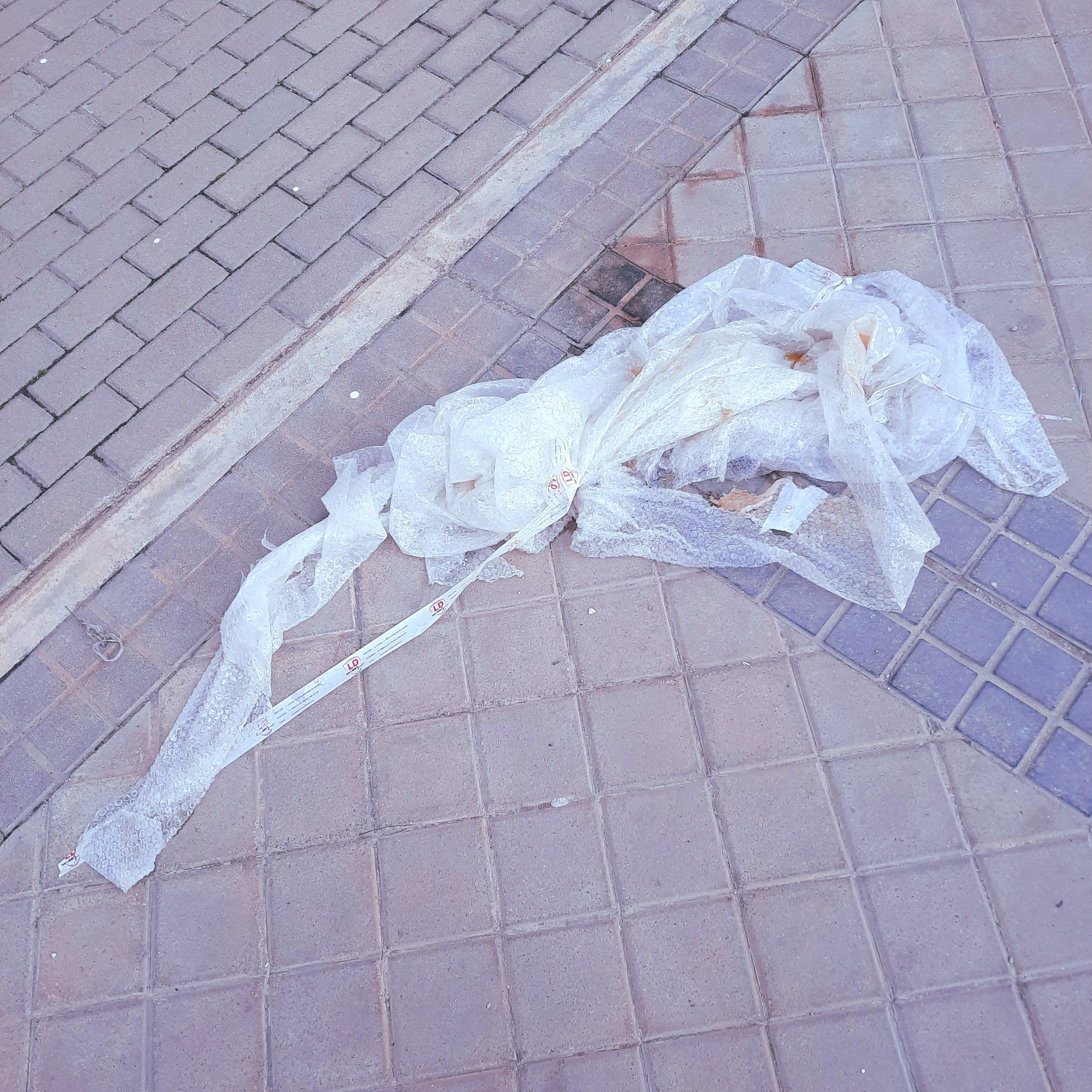 A pile of crumpled plastic shopping bags on a brick sidewalk next to a curb and a drain.