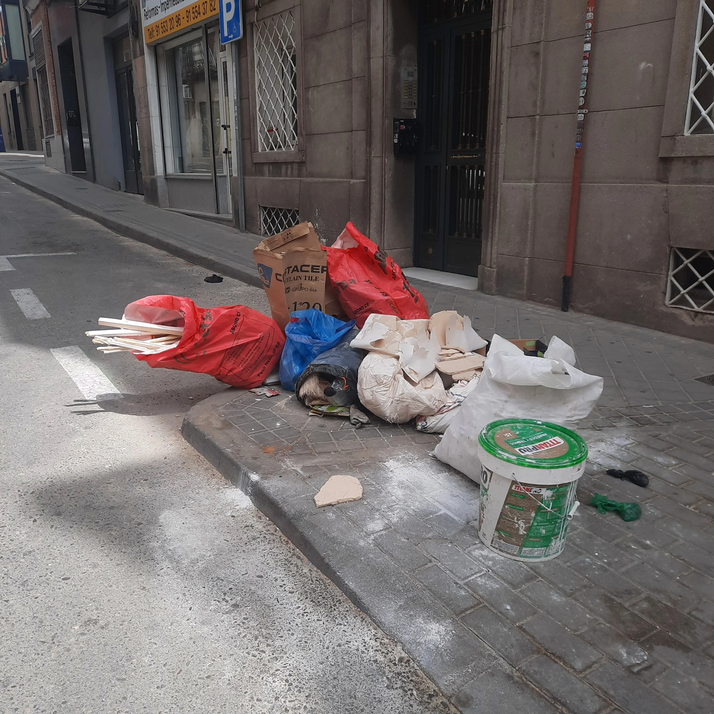 A pile of trash and construction debris including paper bags, a cardboard box, plastic bags, and a large plastic bucket with a green lid on a city sidewalk next to a building with metal security bars and a parking sign.