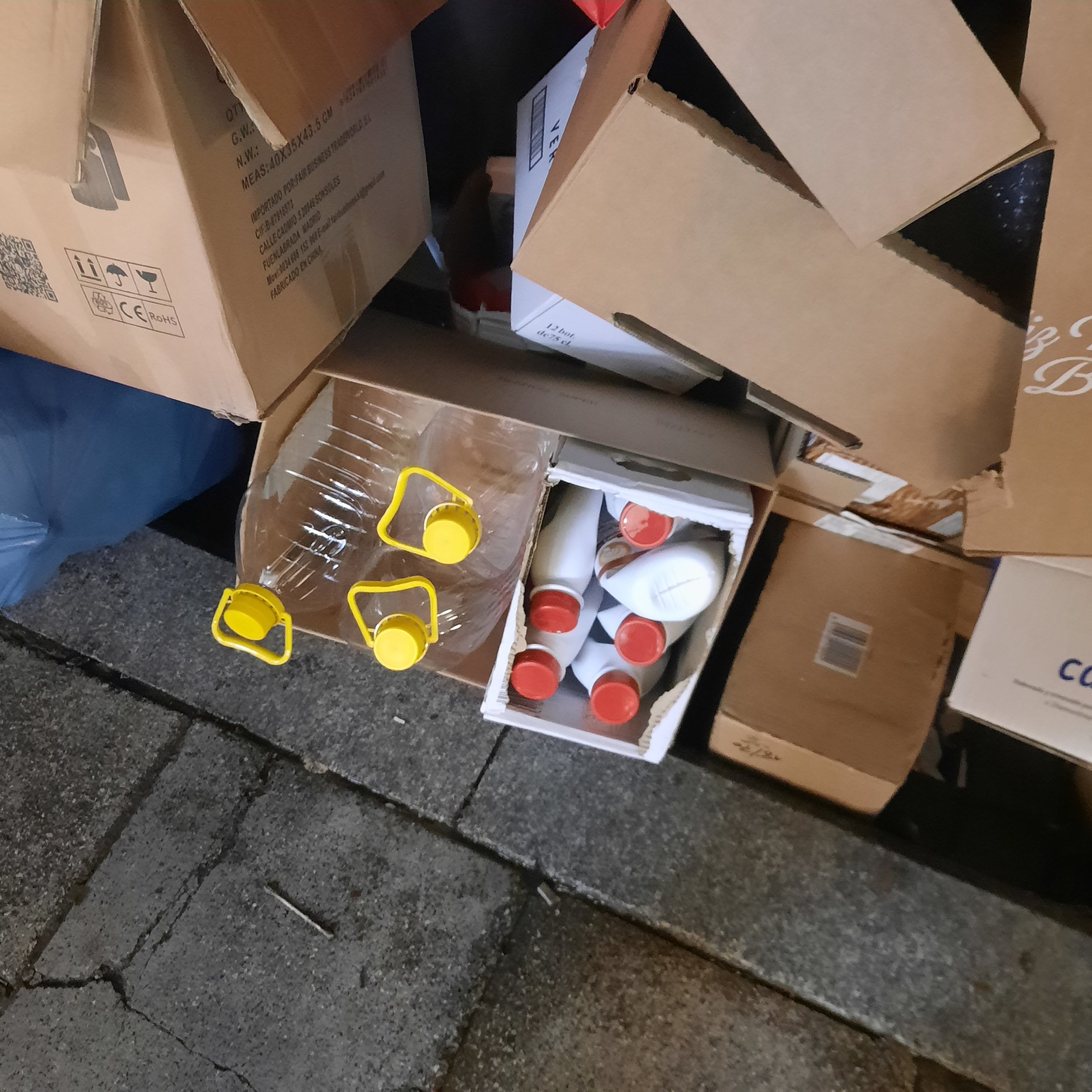 Stacks of cardboard boxes and plastic containers stacked on a wooden pallet and shelves, with some yellow plastic handles and red-capped bottles visible.