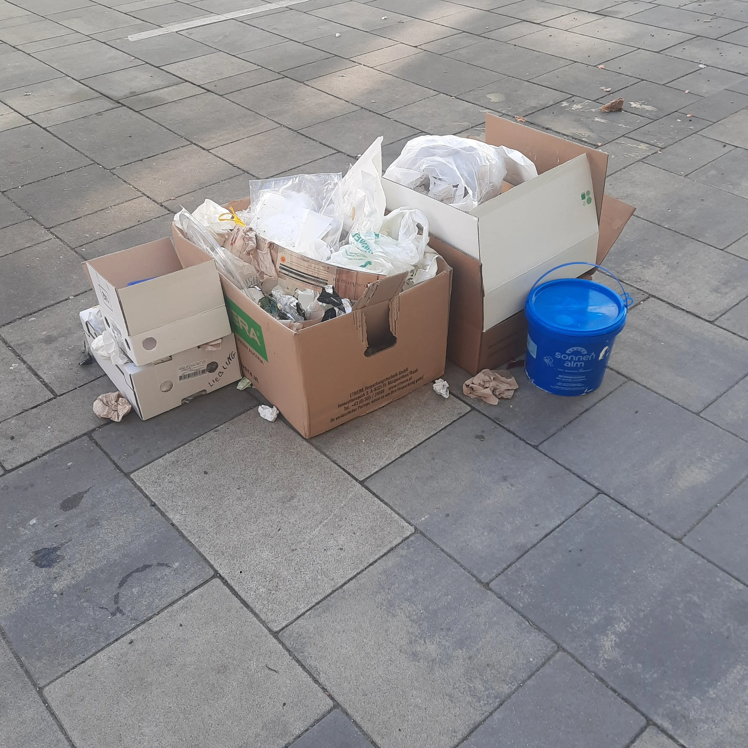 A cardboard box filled with crumpled paper and litter sits on a sidewalk paved with gray bricks, with some additional trash and a blue bucket nearby.