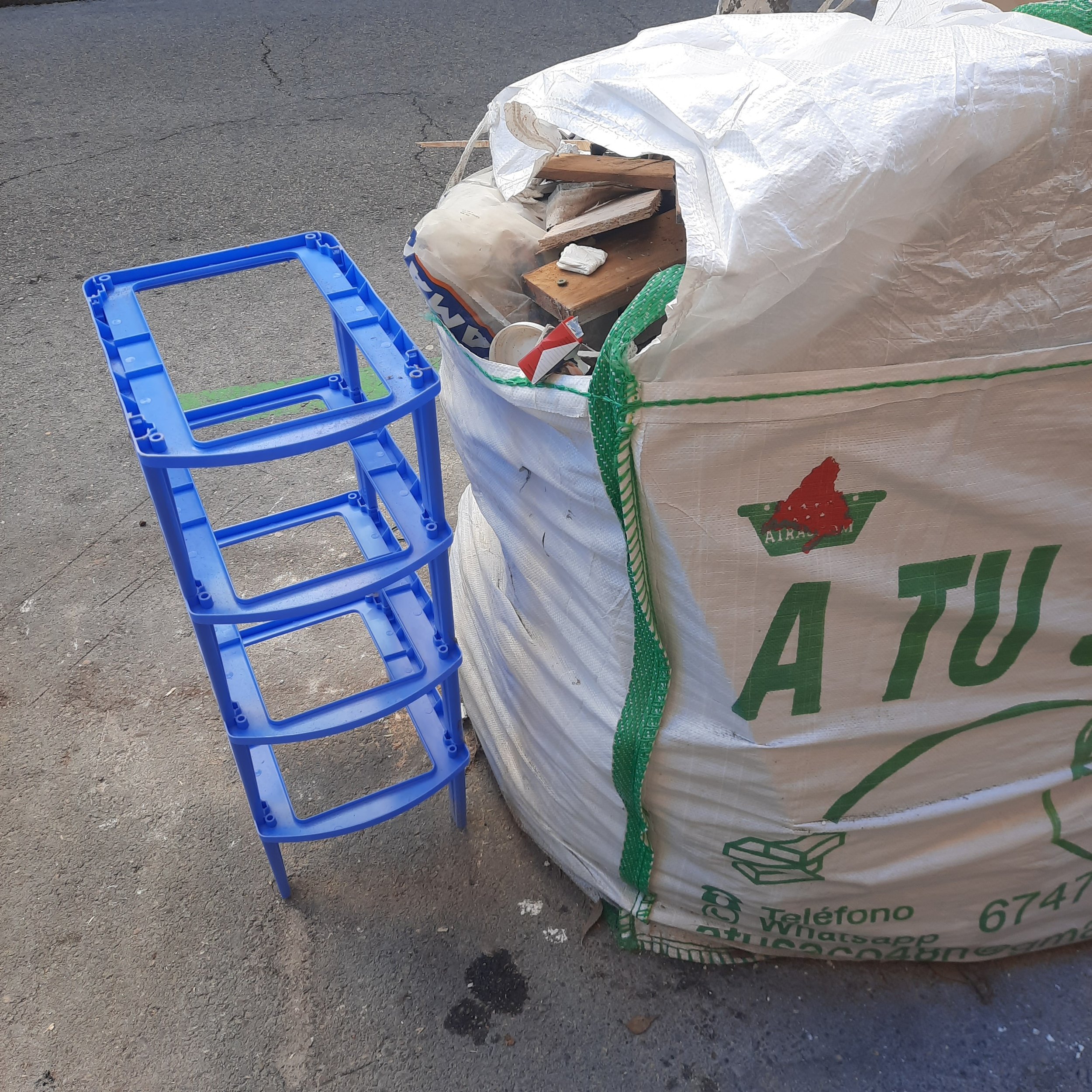 A large white sack filled with assorted wood pieces and debris on an asphalt surface, with a lightweight blue plastic stool beside it.