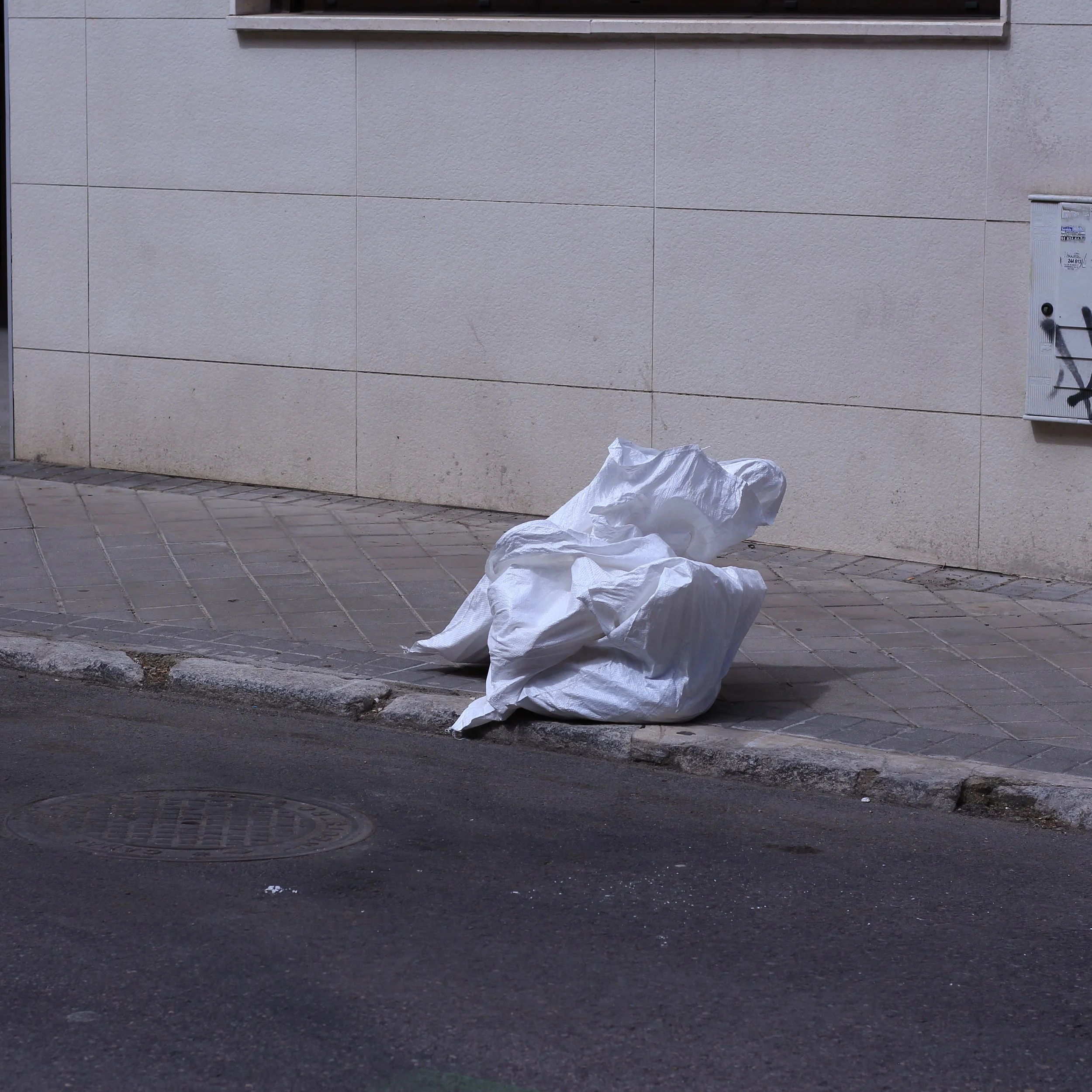 A sidewalk with a large crumpled white plastic bag in front of a beige tiled building wall. There is a small round metal manhole cover and some graffiti on a metallic utility box on the wall.