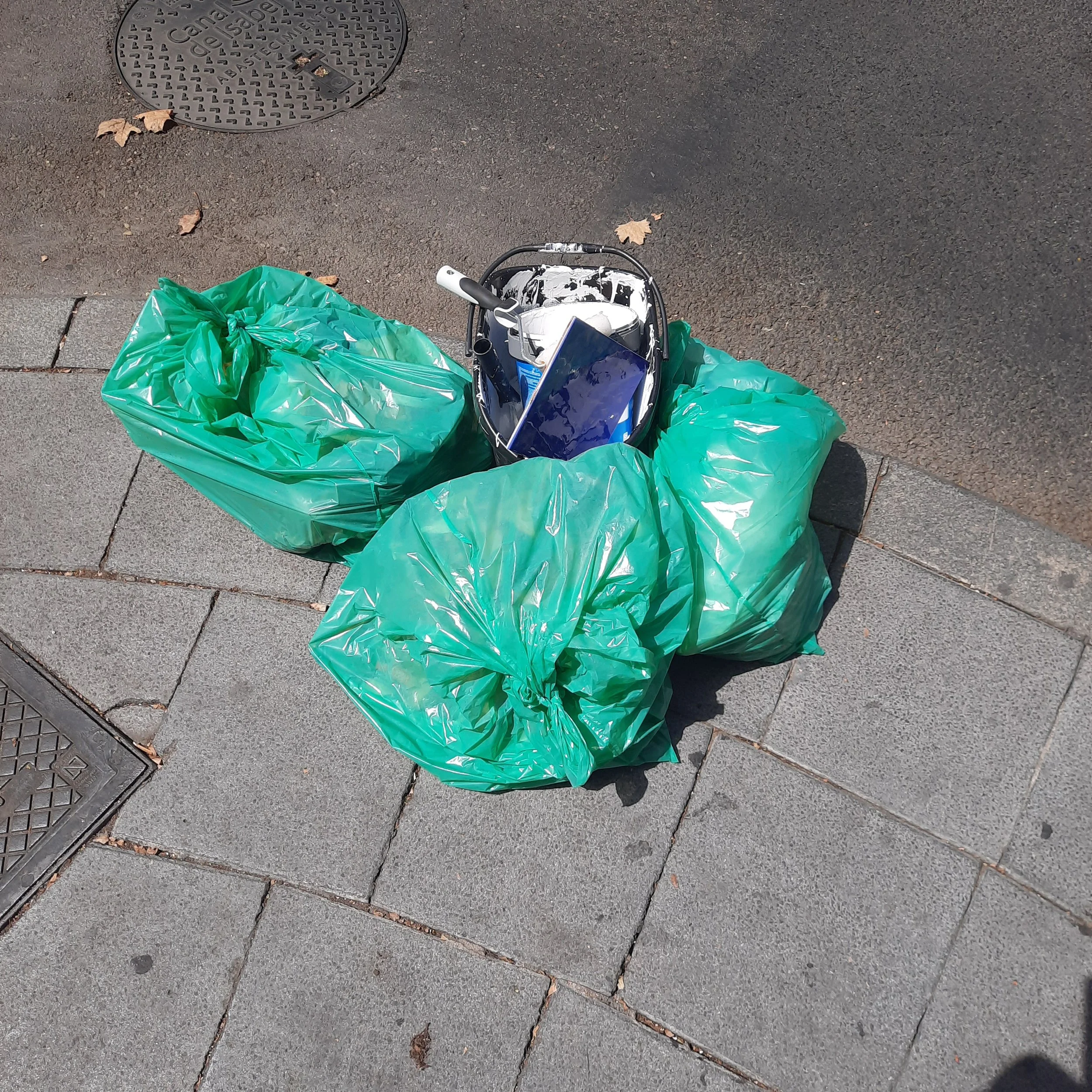A trash bag sits on a city sidewalk near a manhole cover, with a paint roller and paint bucket next to it, and dry leaves scattered on the pavement.