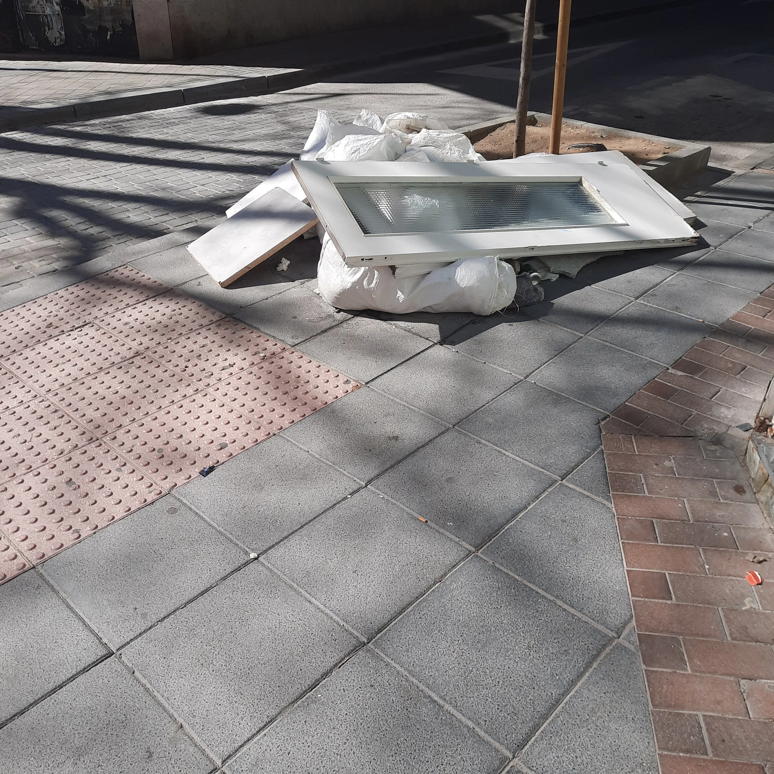 Construction debris including a window frame, white plastic bags, and wooden stakes on a sidewalk.