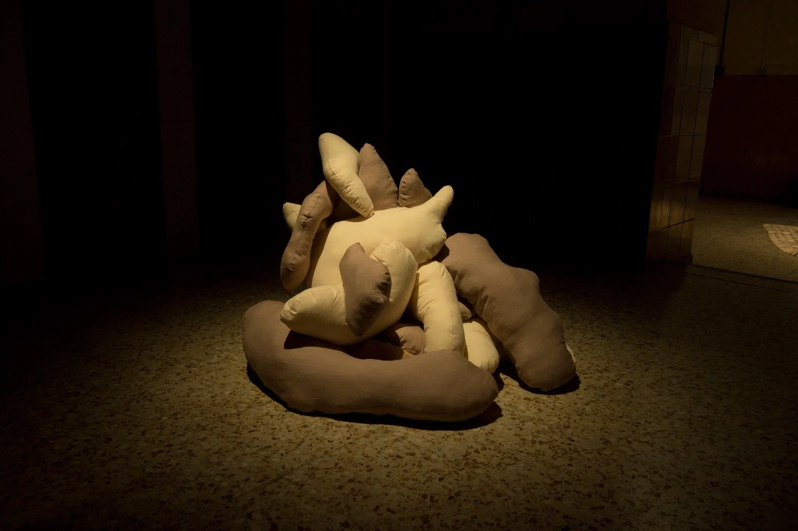 A sculpture of intertwined beige and light brown fabric forms, resembling abstract human figures, positioned on a speckled grey floor with dramatic spotlight lighting.