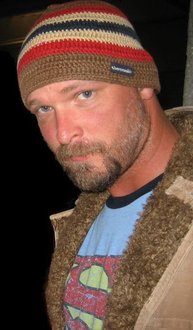 A man with a light beard and mustache wearing a colorful striped beanie, a blue T-shirt with a Superman logo, and a brown furry jacket.