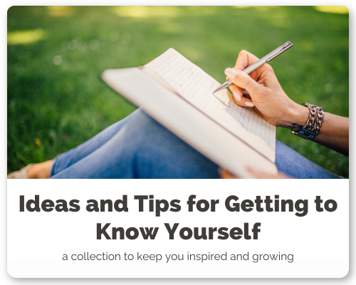 A person sitting outdoors on grass, writing in a notebook with a pen, wearing jeans and a wristwatch, with a book resting on their lap.