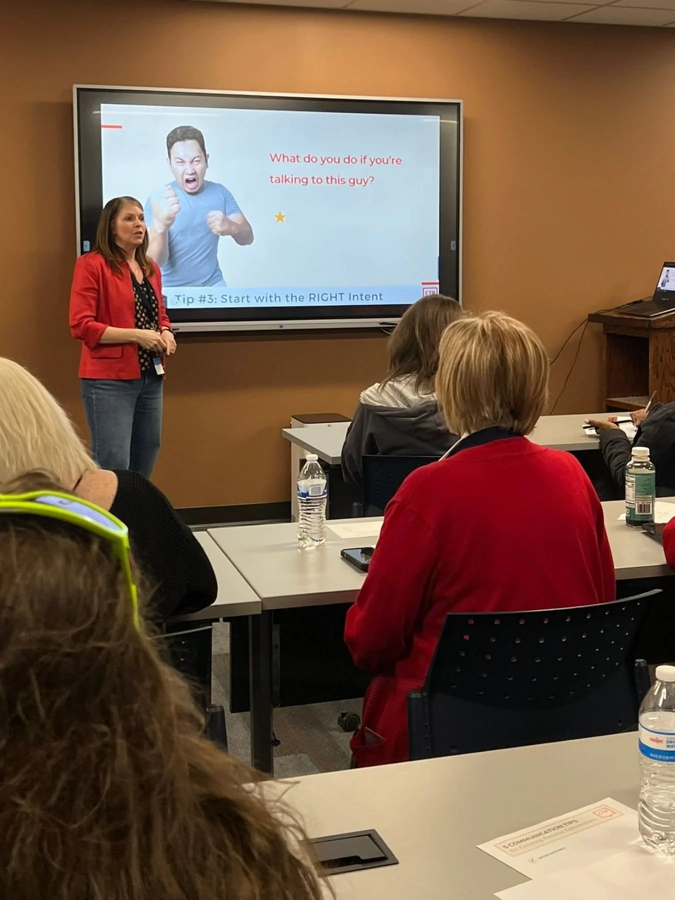 A woman in a red blazer and patterned top giving a presentation in a classroom, with a slide on a TV screen behind her asking 'What do you do if you're talking to this guy?' The slide shows a man with an angry expression making a fists gesture. Students are seated at desks, some with water bottles.