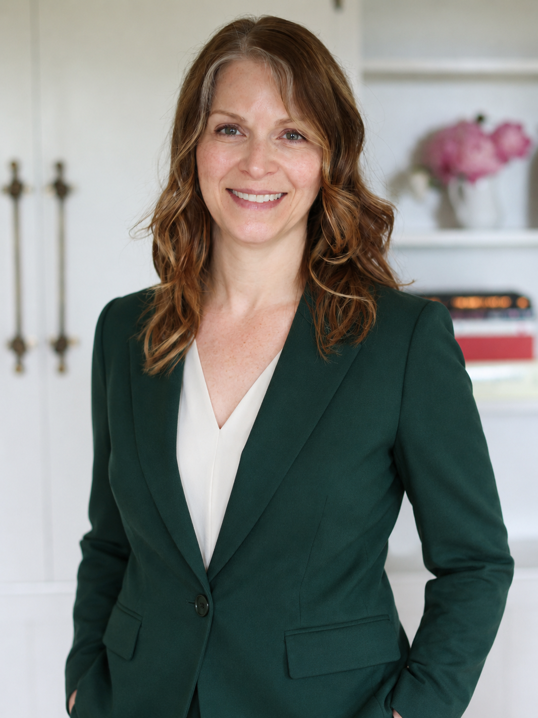 A woman with red hair smiling, wearing a dark green blazer and white blouse, standing indoors with a white bookshelf and pink flowers in the background.