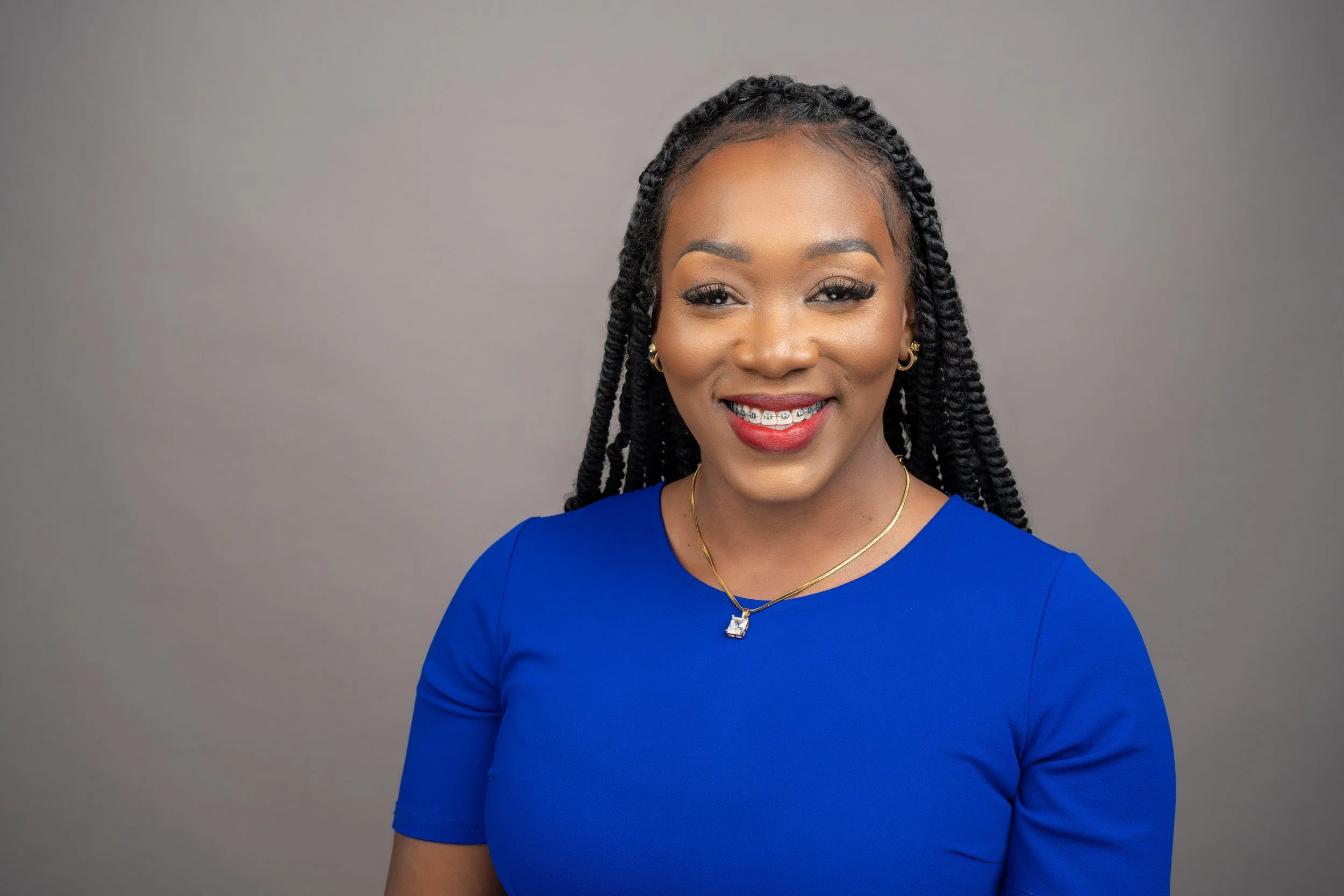 A smiling woman with braided hair wearing a blue dress, red lipstick, and jewelry, against a plain gray background.