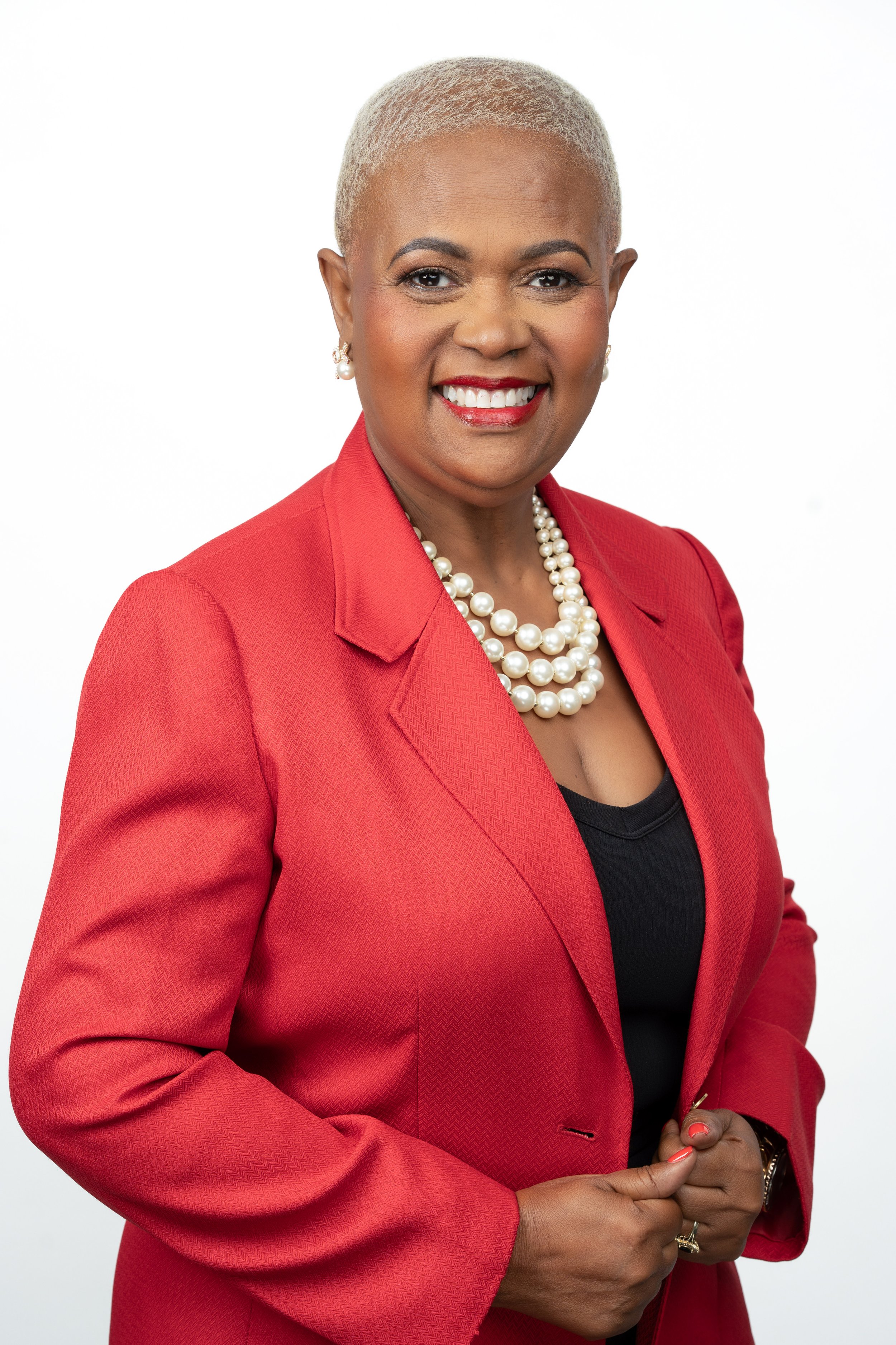 A confident African American woman with very short, platinum blonde hair, wearing a red blazer, pearl necklace, pearl earrings, and red lipstick, standing against a plain white background.
