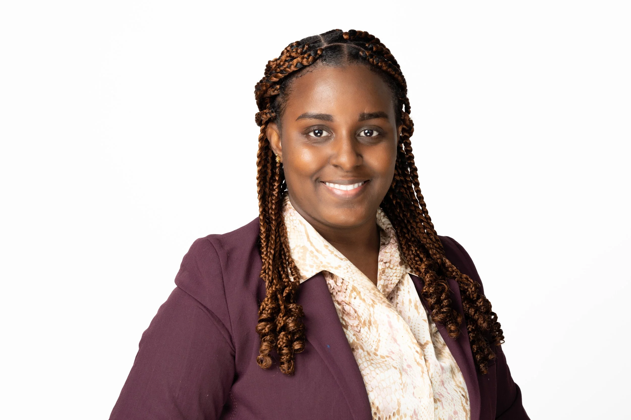 A woman with brown skin and long braided hair wearing a purple blazer, beige patterned blouse, and gold earrings, smiling against a white background.